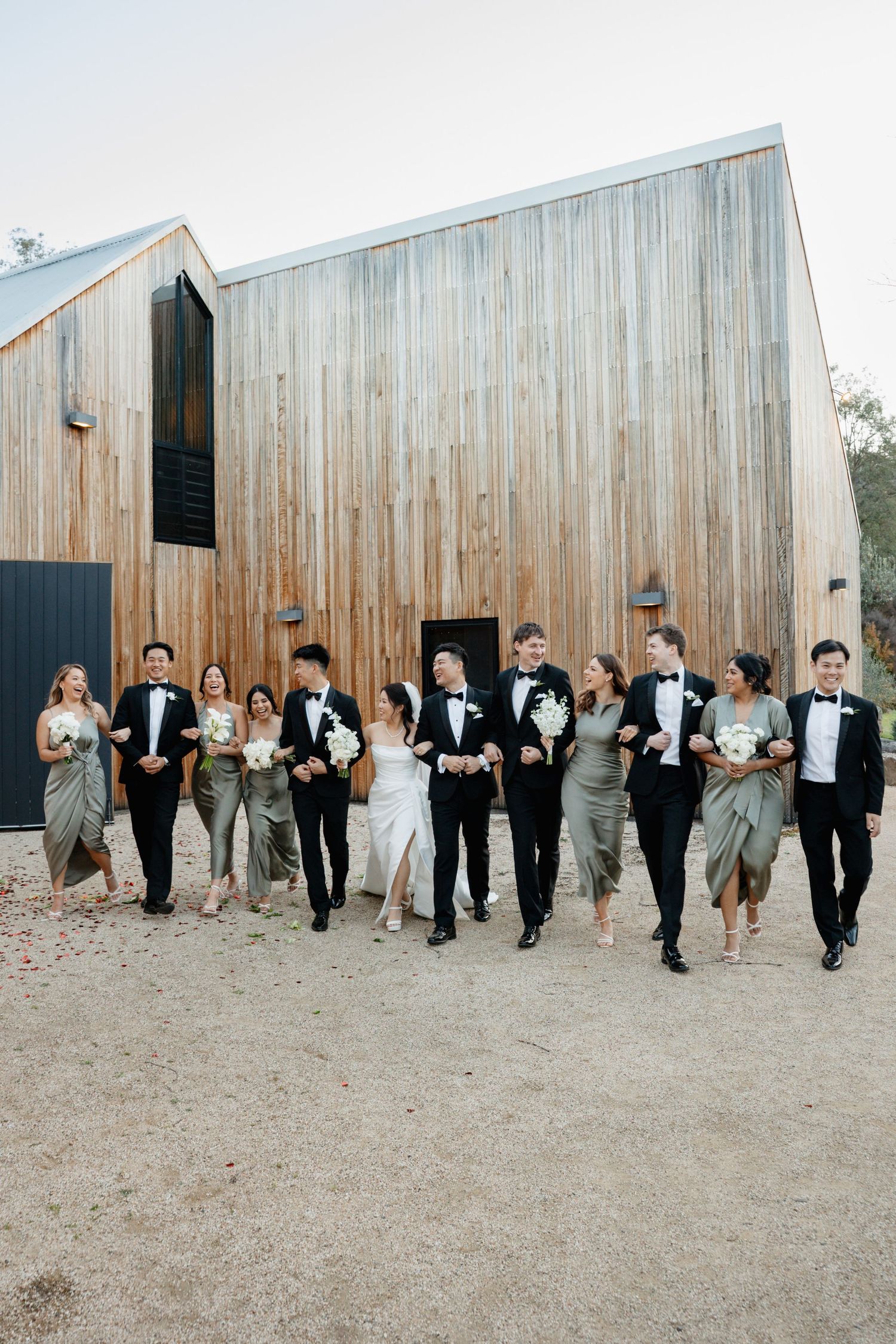 Wedding party walks together in front of rustic wooden barn structure.