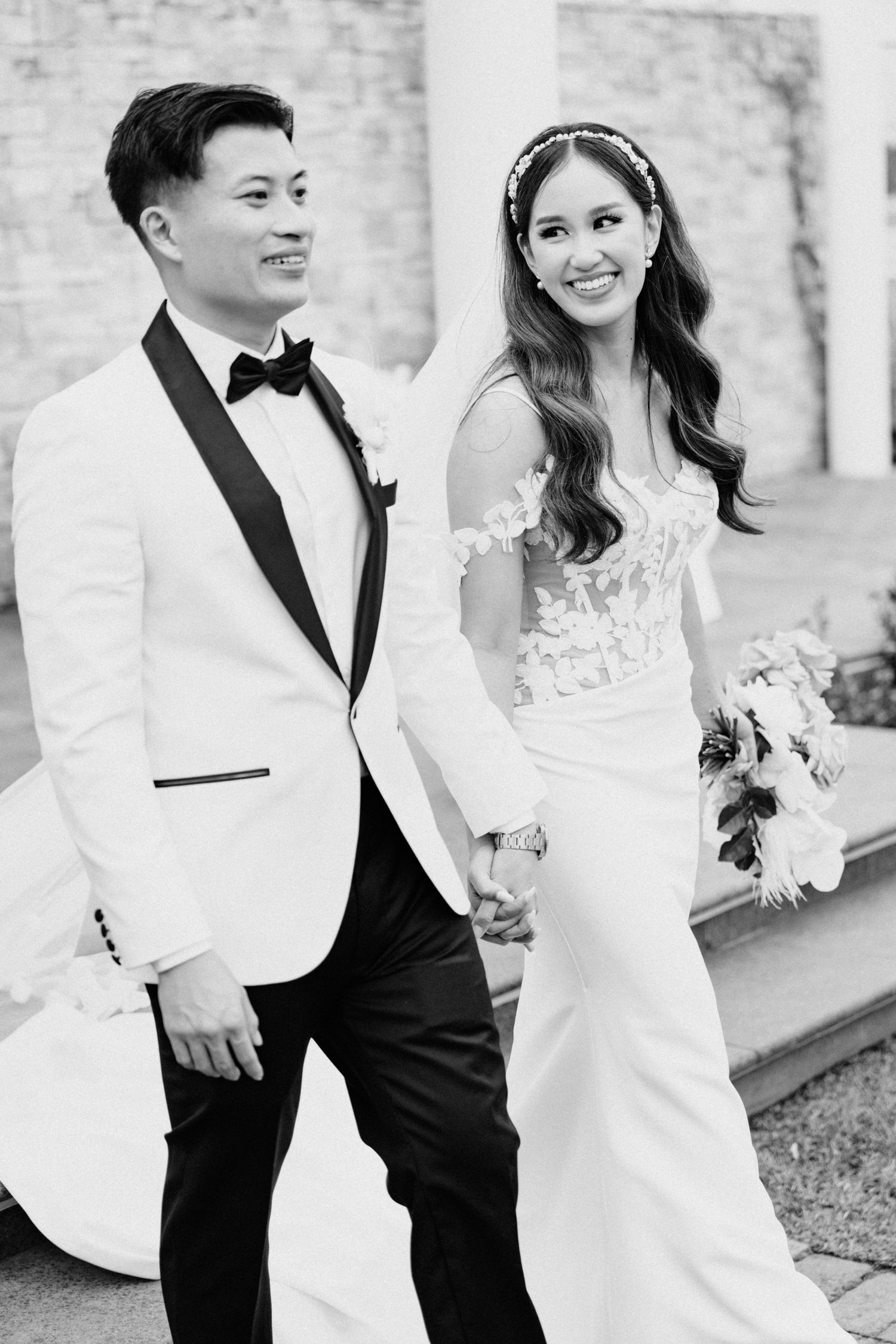 A bride in a lace gown and groom in white tuxedo jacket pose together for elegant black and white portrait.