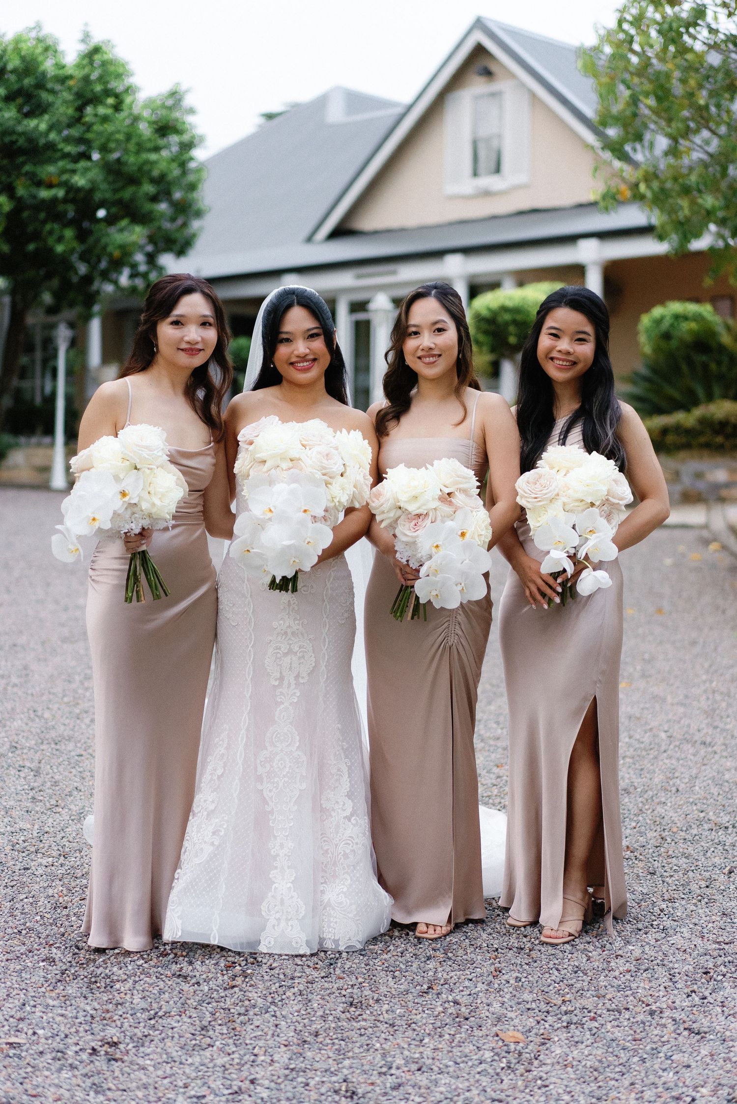 Bridal party in neutral tones holding white bouquets pose in front of house.