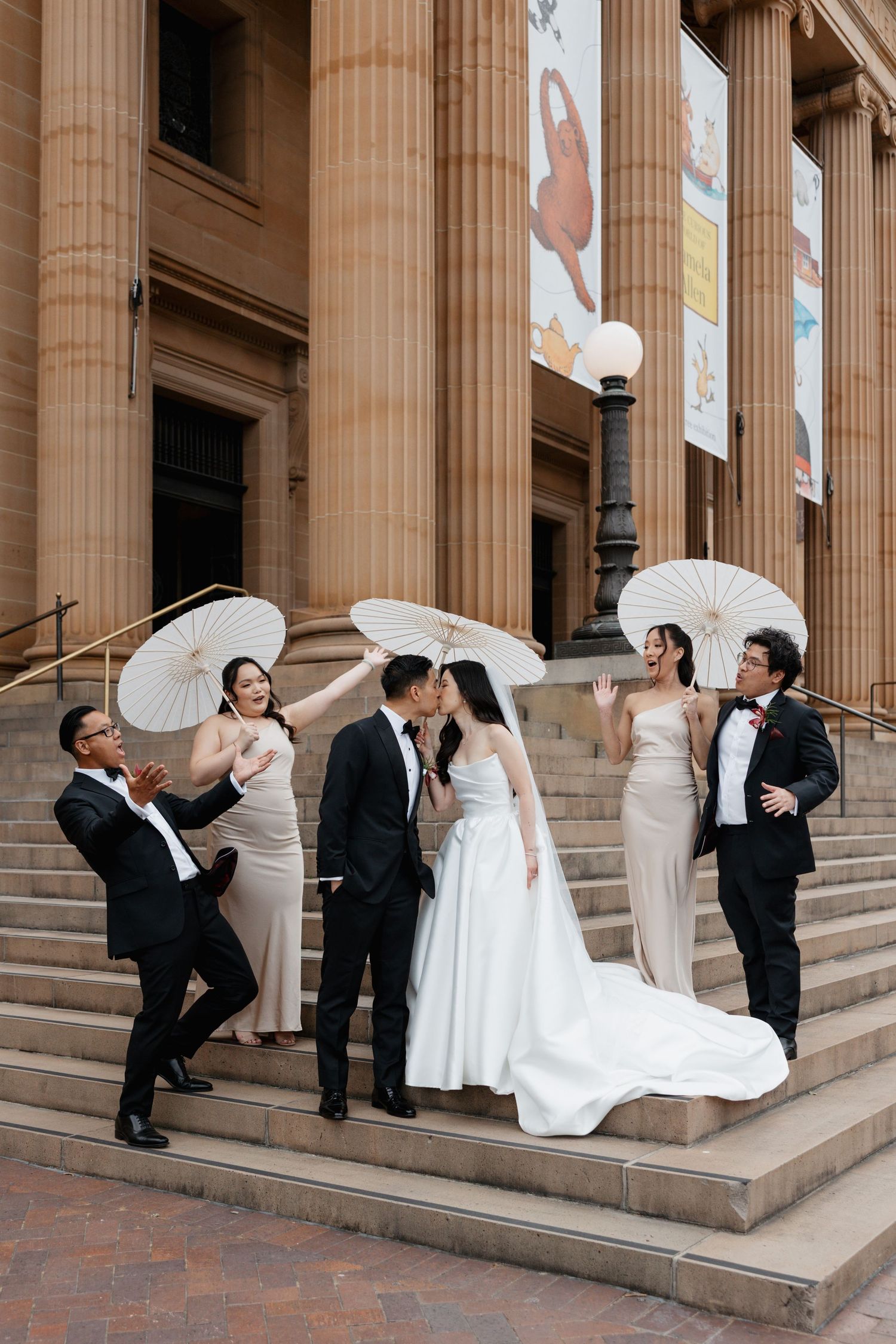 Wedding party poses with white umbrellas on grand stone steps with classical columns.