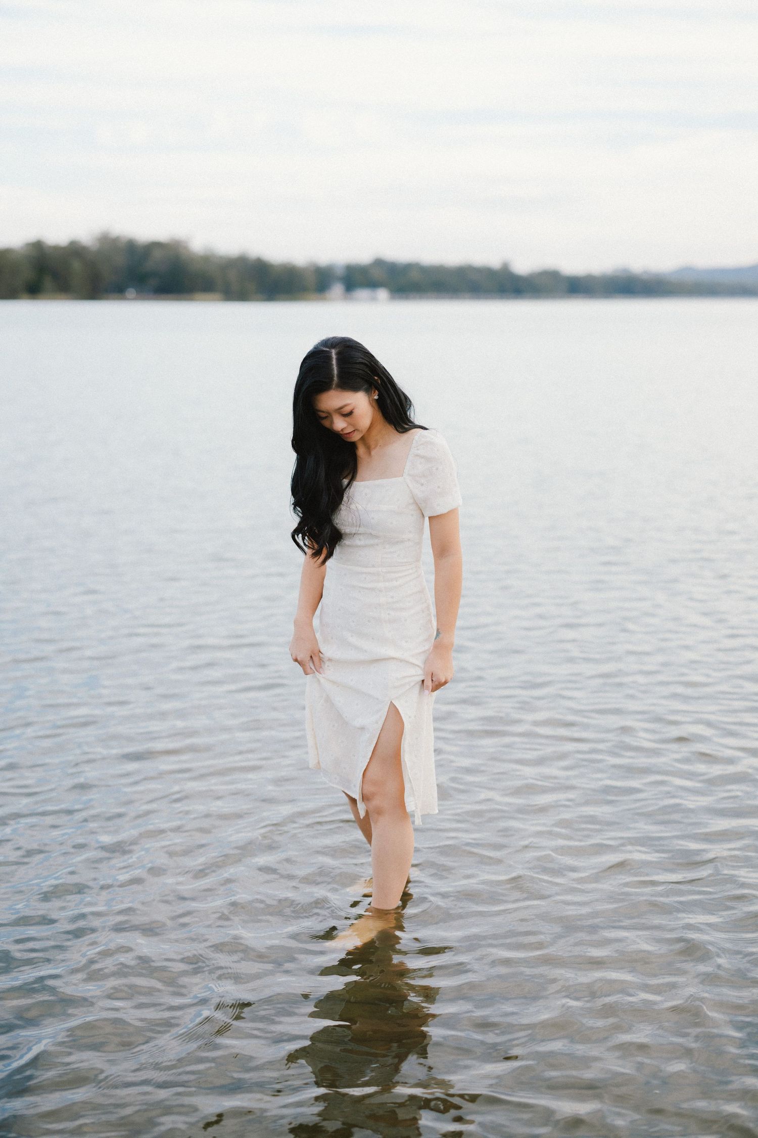 A person in a white dress wades through tranquil lake waters on a cloudy day.