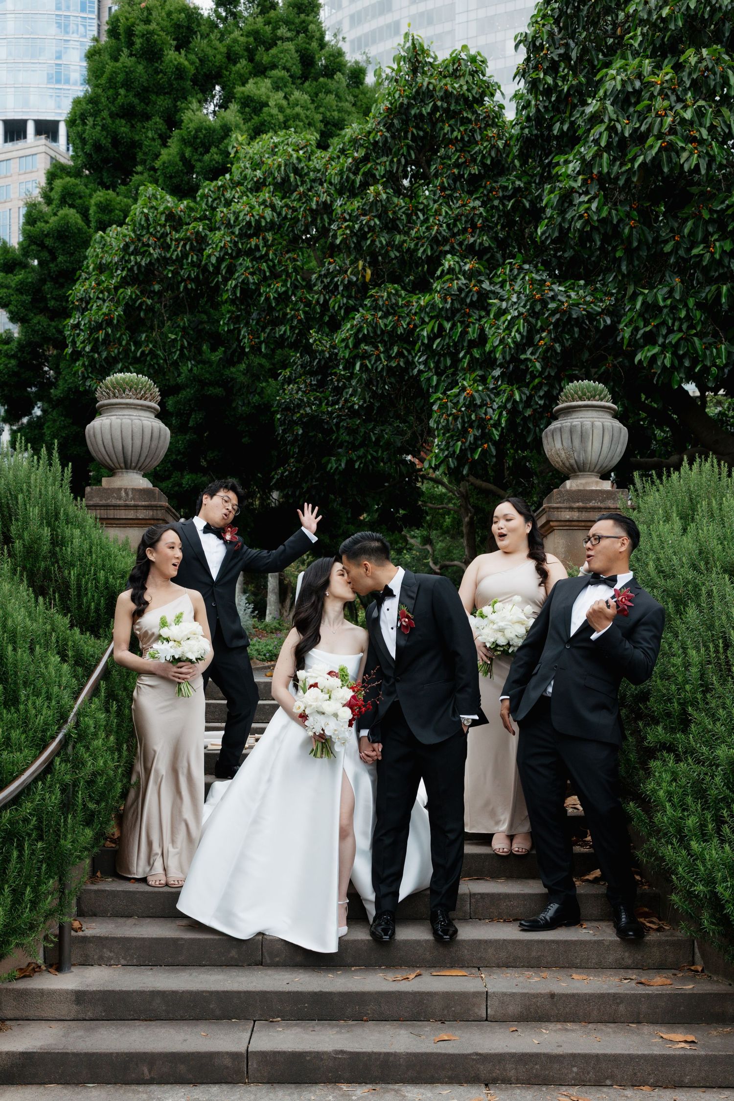 Wedding party poses in sequence on stone steps with decorative pillars in a garden setting.