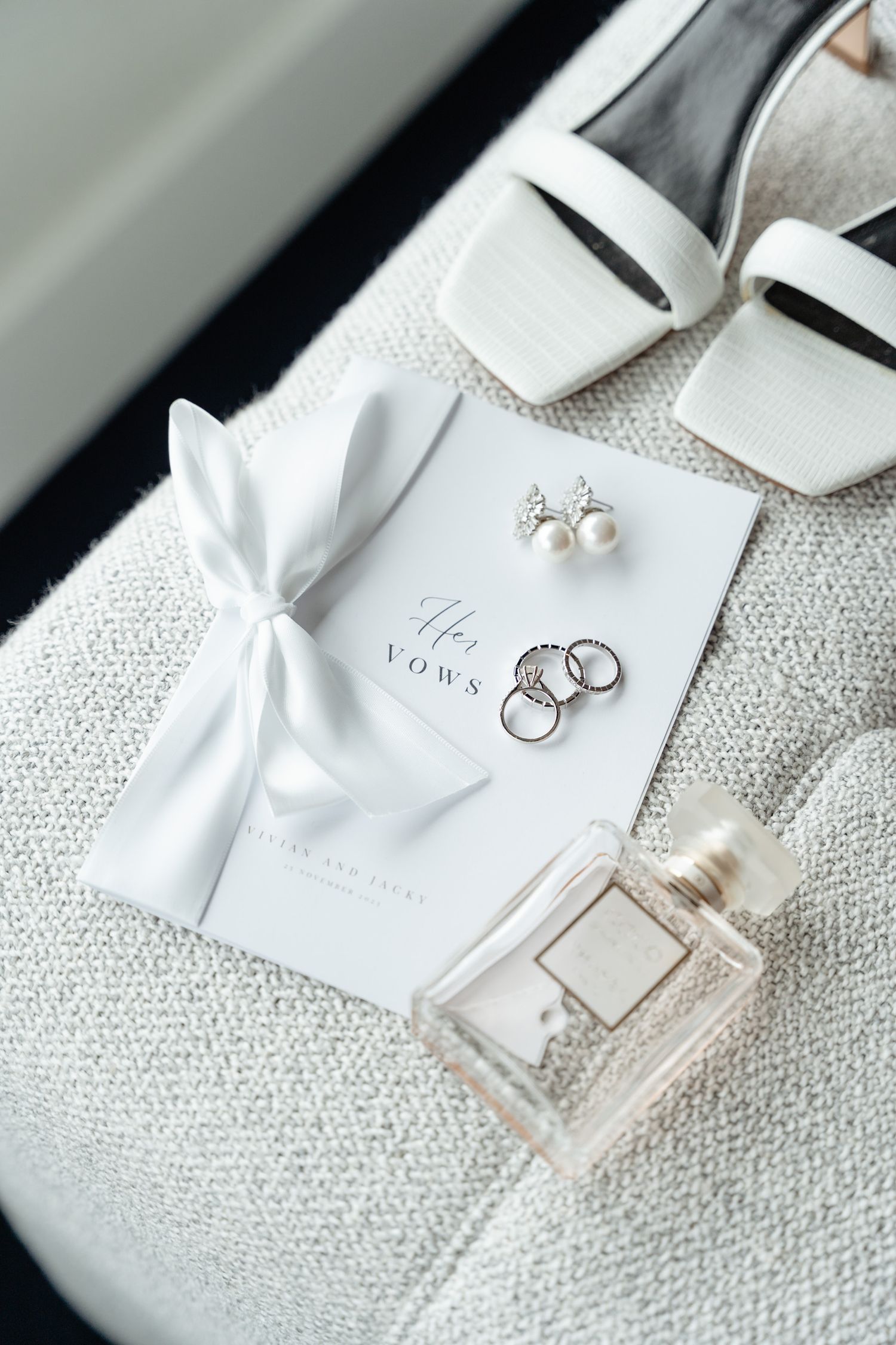 Wedding accessories including perfume, rings, and white bow details arranged on textured white surface.