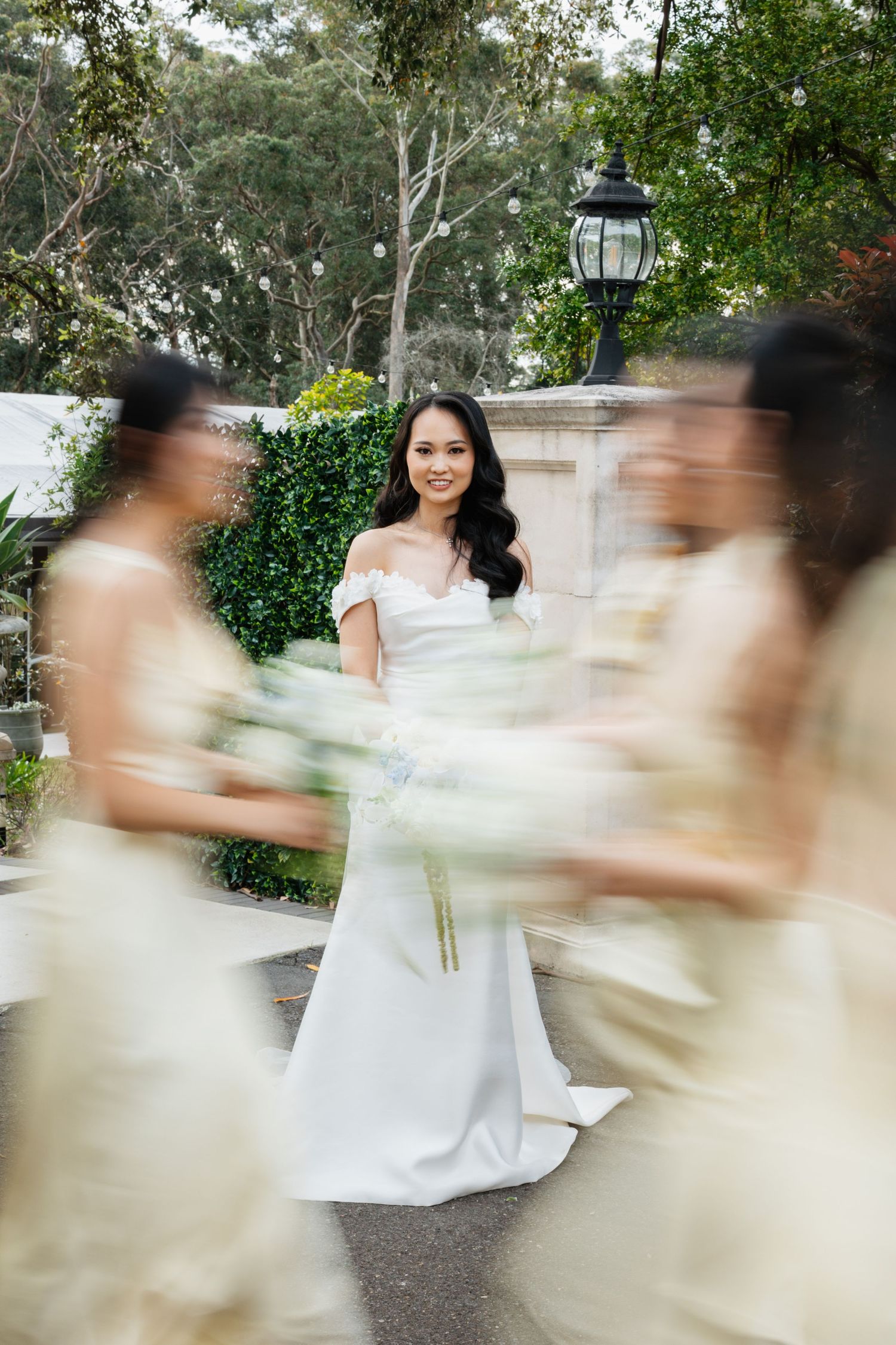 Artistic motion blur photo of bride in white dress standing in garden setting with lamp post.