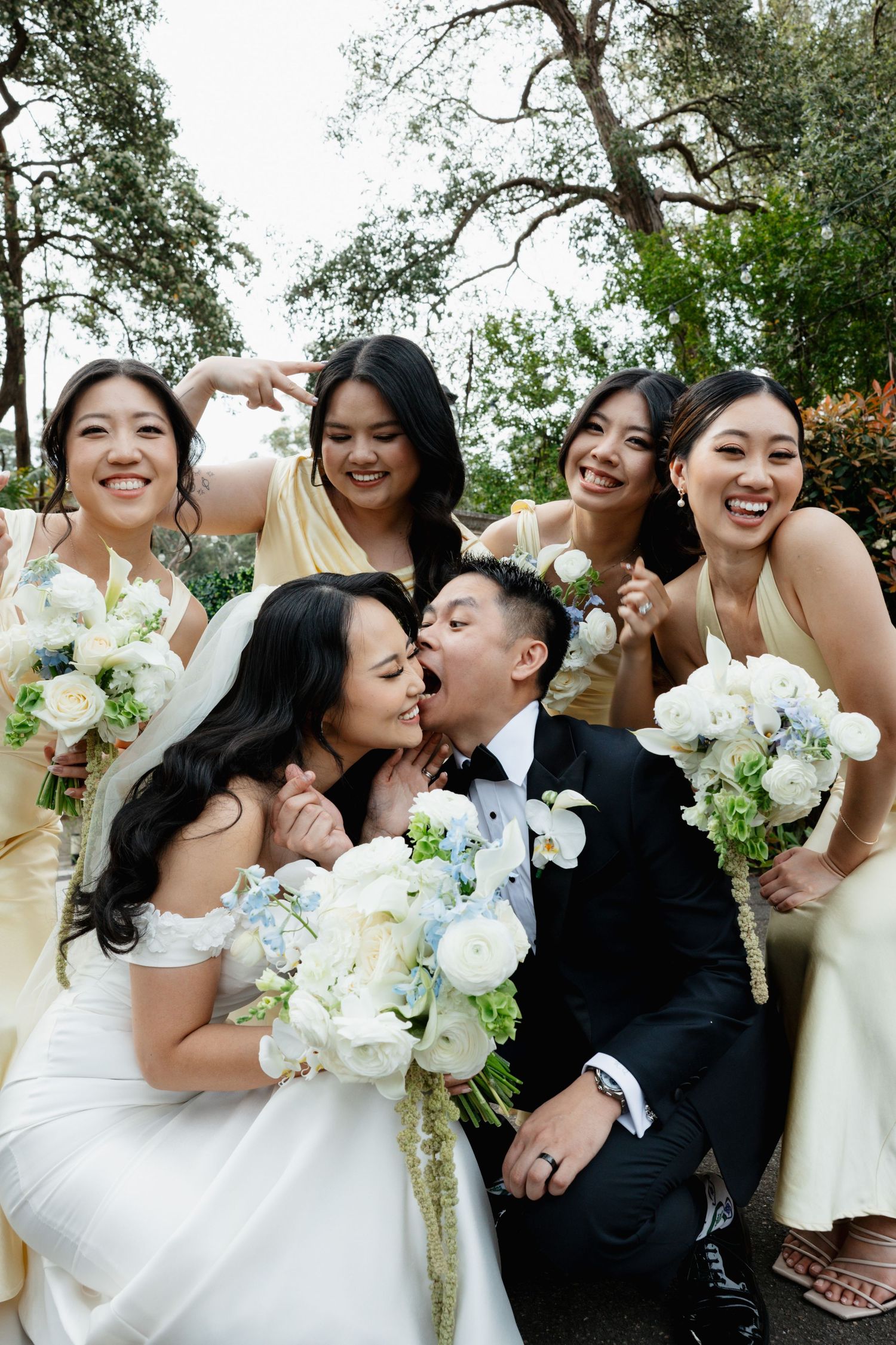 Wedding party poses with white floral bouquets against natural outdoor backdrop.
