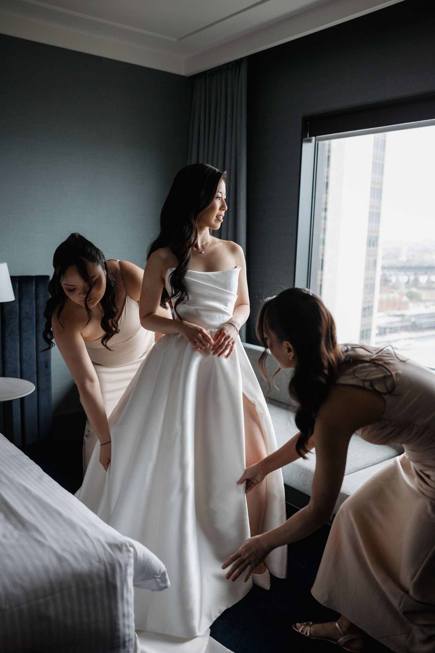 Bridesmaids in blush dresses help adjust a white wedding gown in a hotel room with city views.
