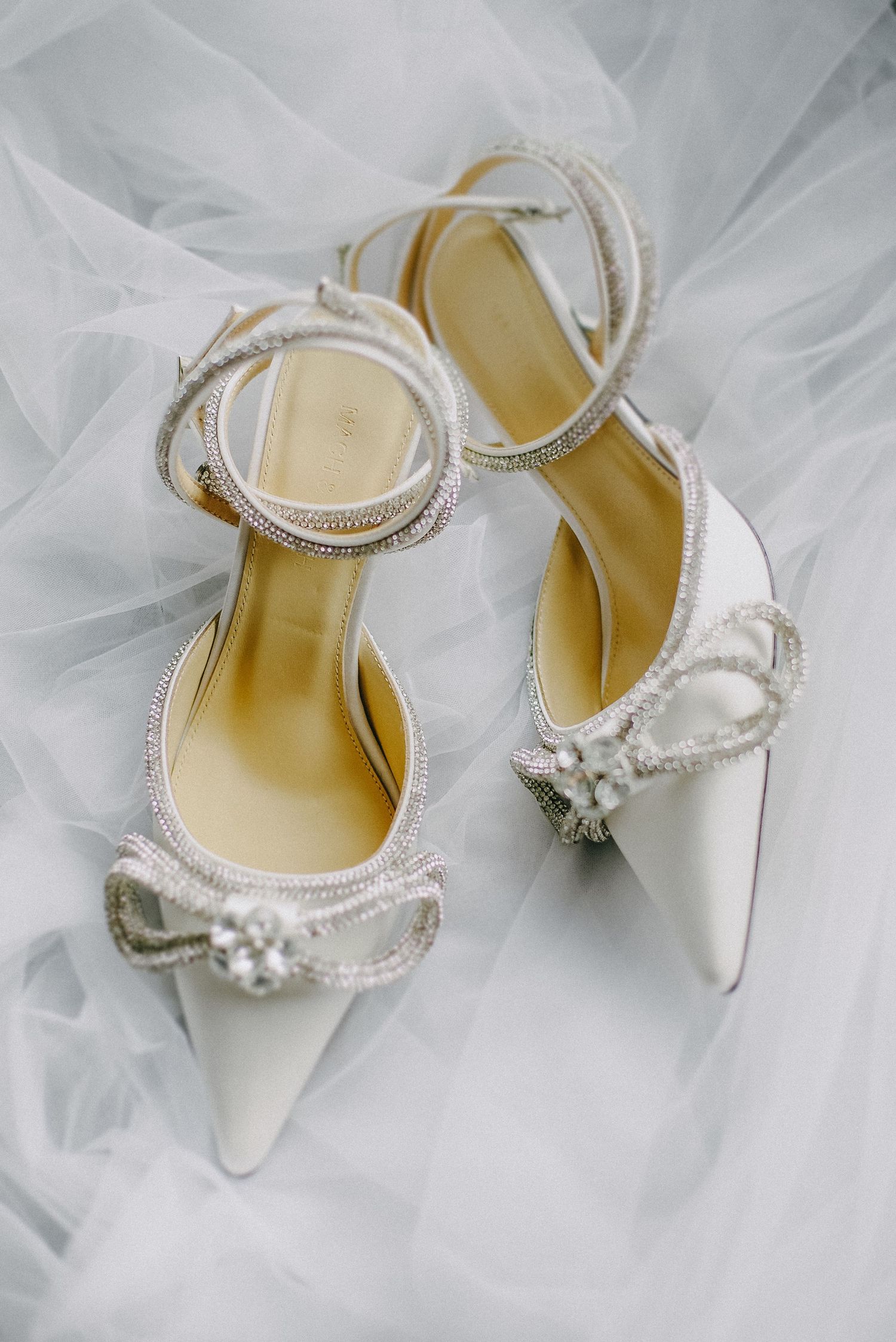 Elegant white bridal shoes with crystal embellishments and delicate straps displayed on tulle fabric.