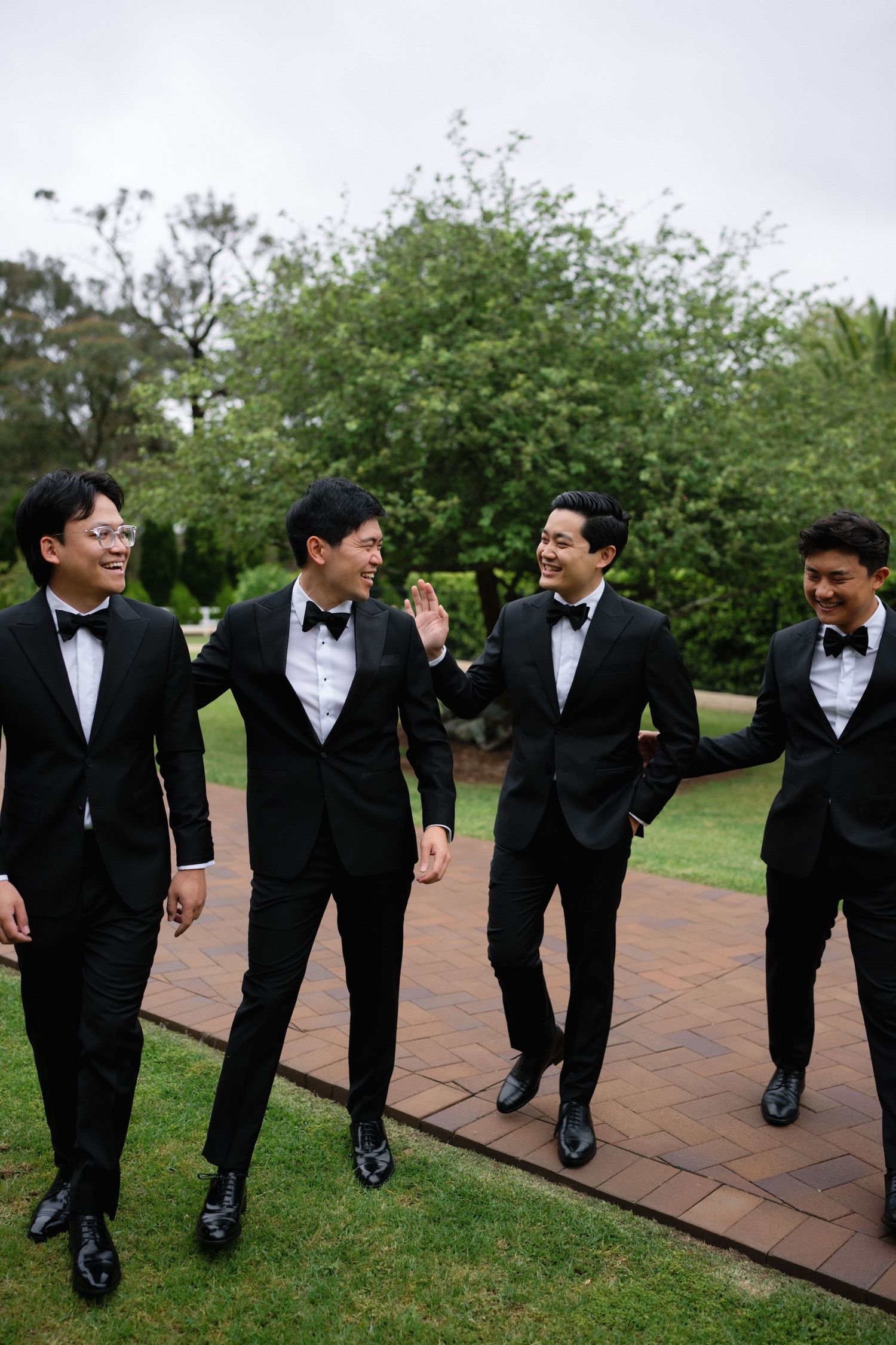 A group of groomsmen in black tuxedos laugh together while walking on a brick pathway outdoors.