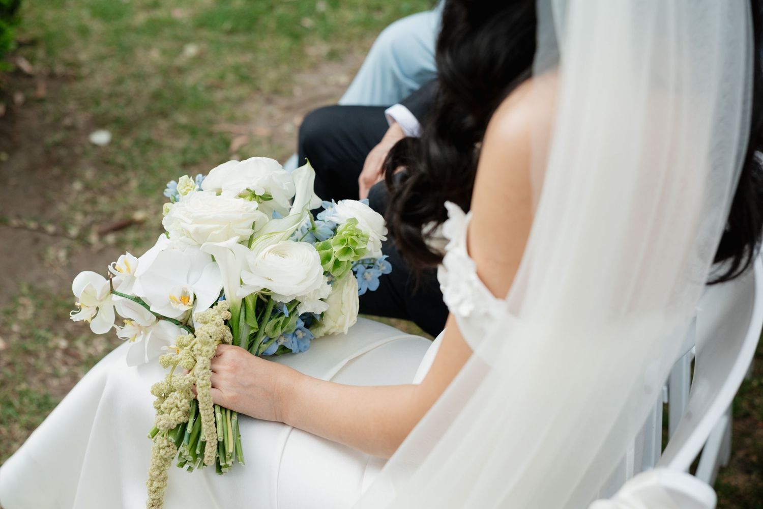 A delicate white bridal bouquet with roses and greenery is held against a flowing wedding dress veil.