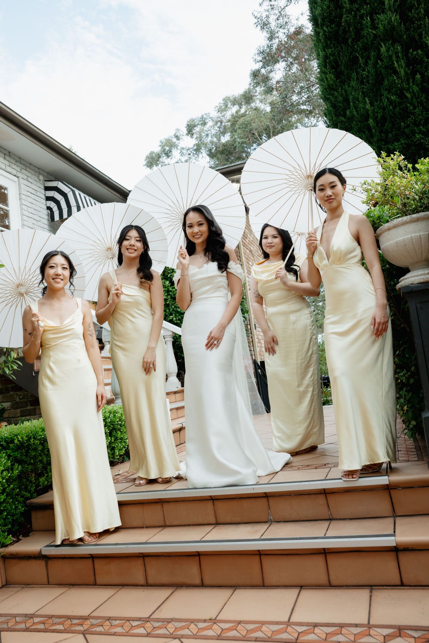 Wedding party in cream dresses poses with parasols on outdoor deck steps.