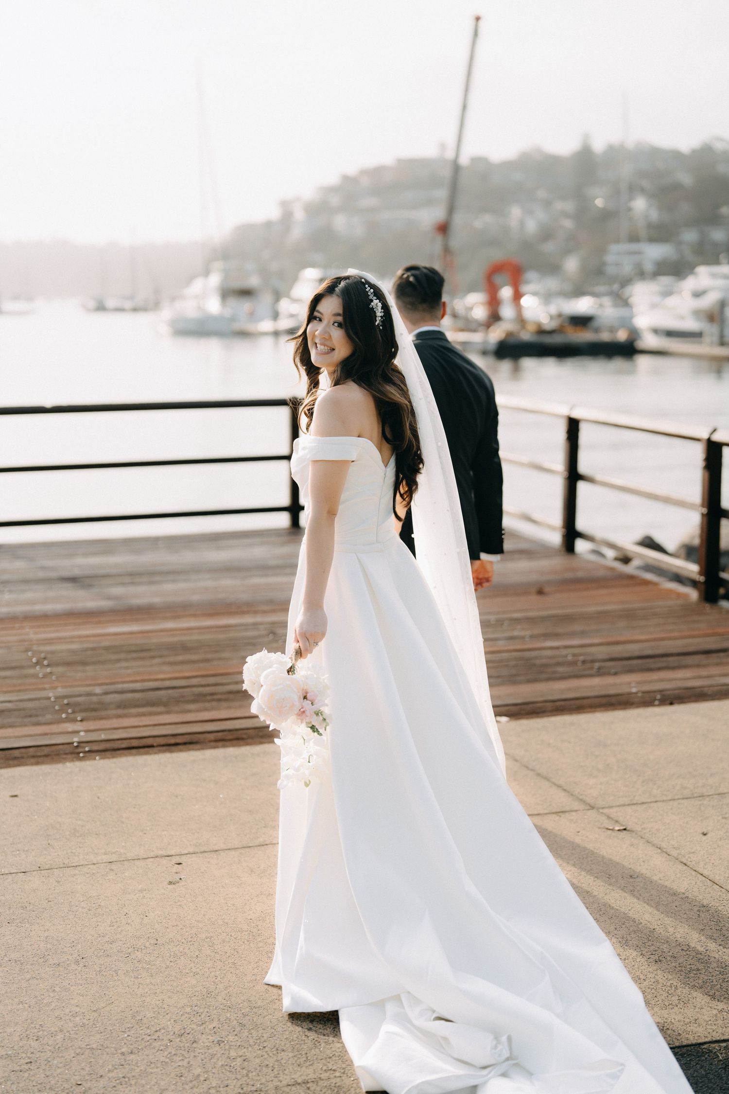 Couple in wedding attire walks along wooden pier with harbor view.