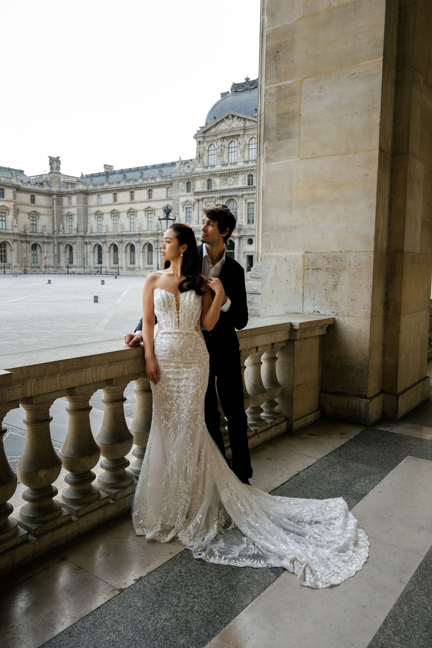Elegant couple shares intimate moments on a grand balcony with classical architecture and stone columns in view.