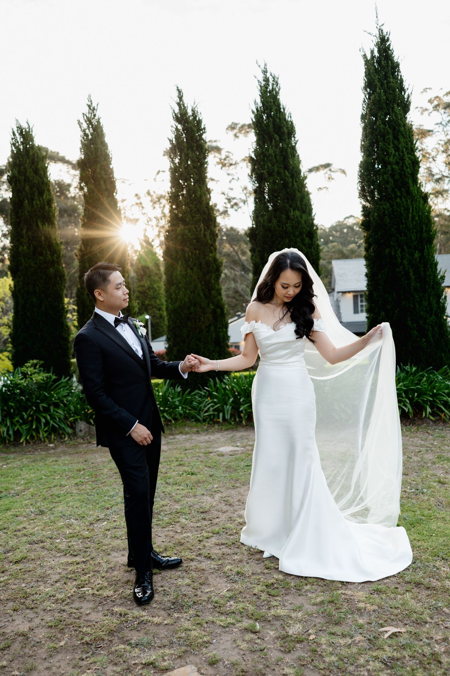 A wedding couple poses in a garden with cypress trees and sunlight, the bride showing off her elegant white dress.