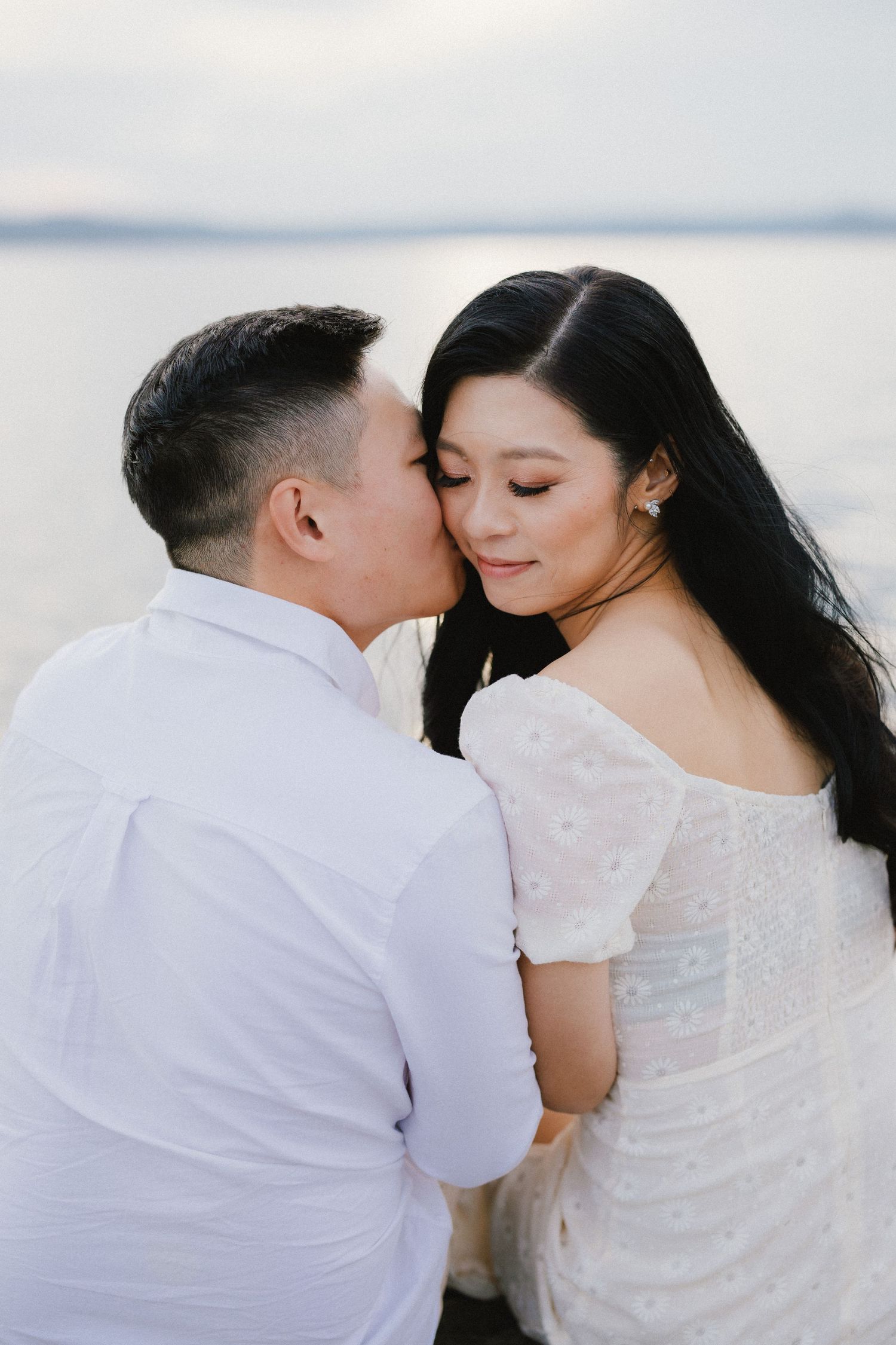 A romantic couple in white embraces tenderly against a serene waterfront backdrop.
