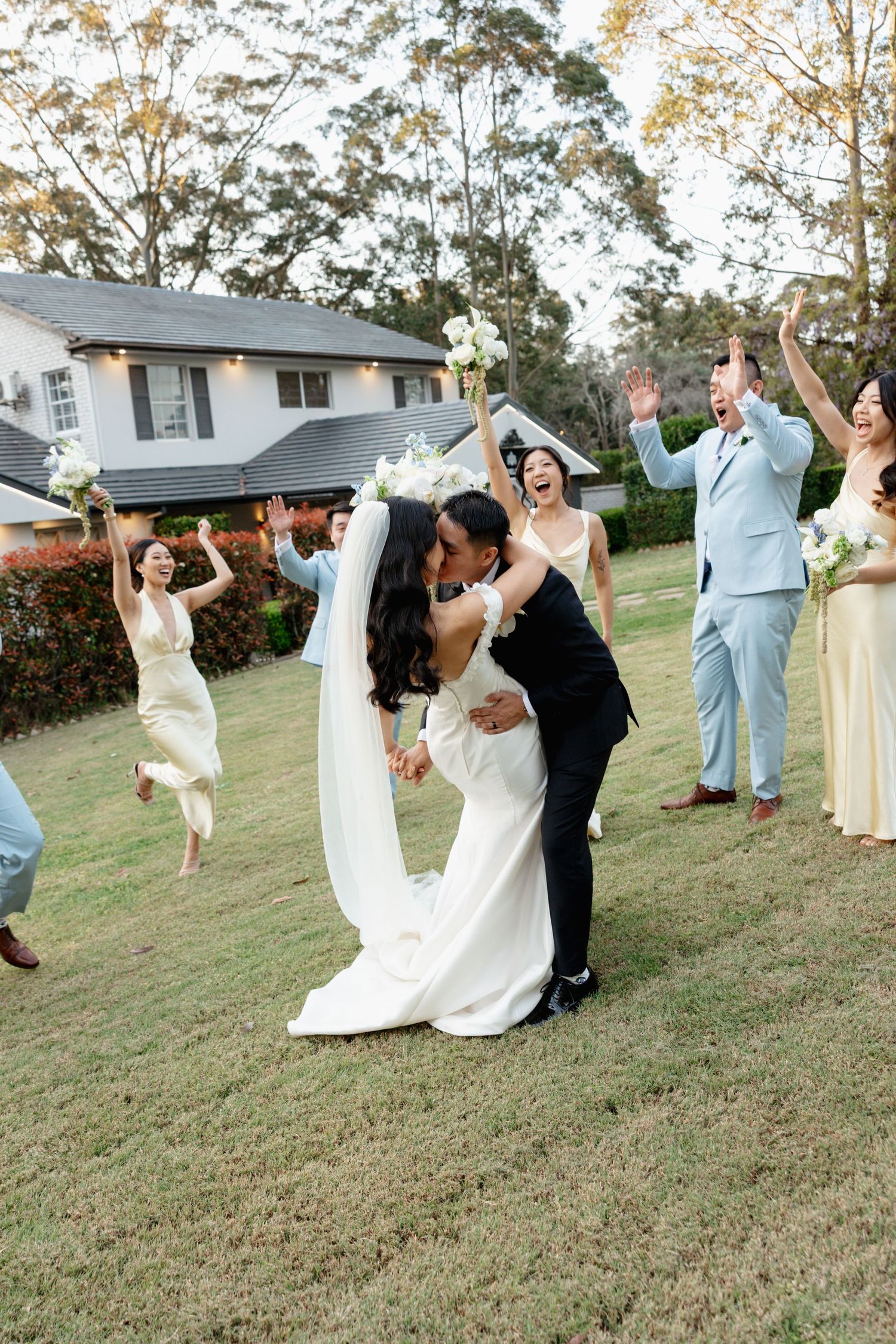 Wedding party poses playfully on a grassy hill outside residential homes.