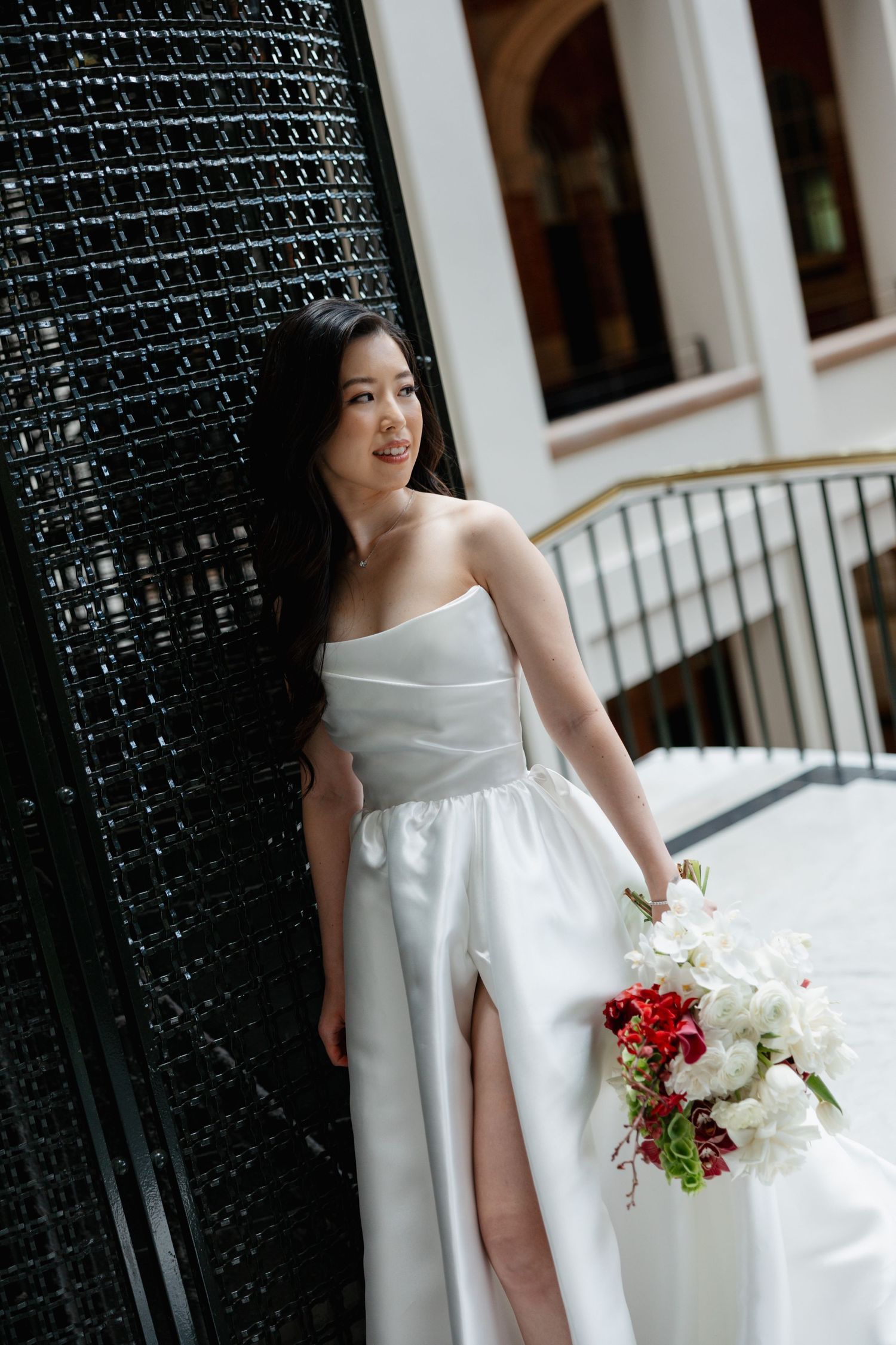 Modern bride in sleek white gown with thigh slit poses against textured wall holding red and white bouquet.