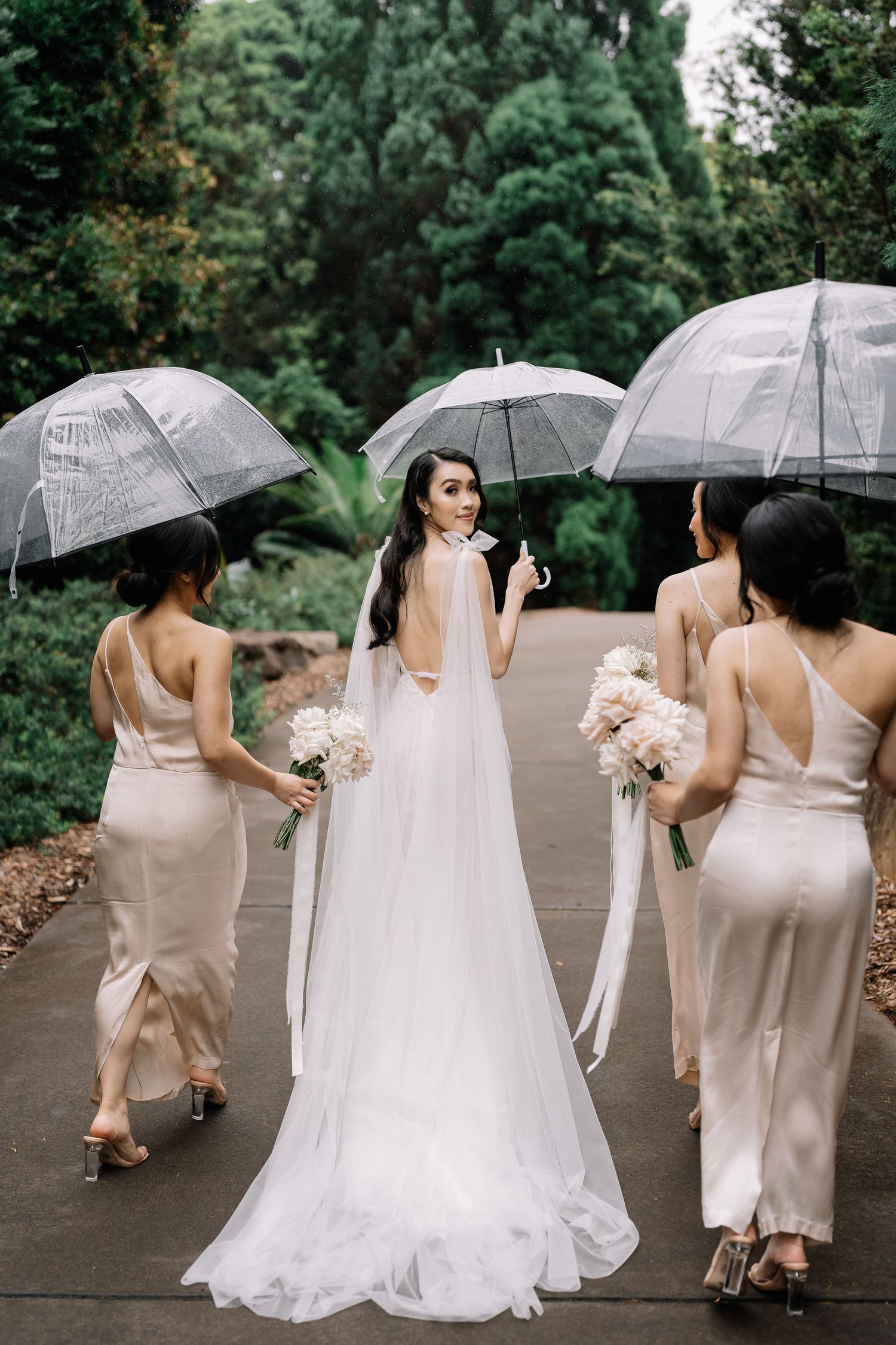 Bridesmaids in champagne dresses hold clear umbrellas while walking with bride.