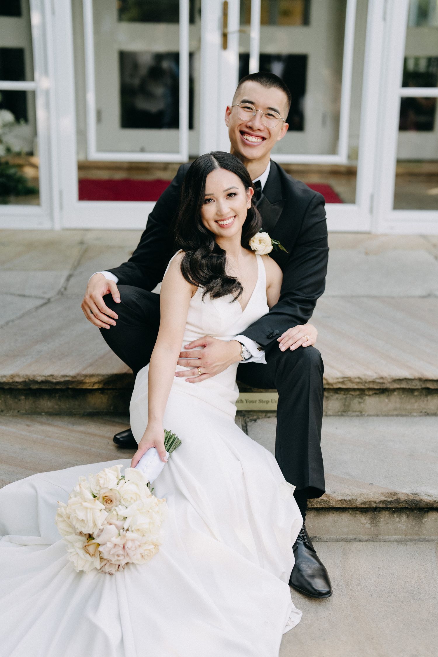 A bride in a white wedding dress sits on stone steps while posing with her groom in a black suit.