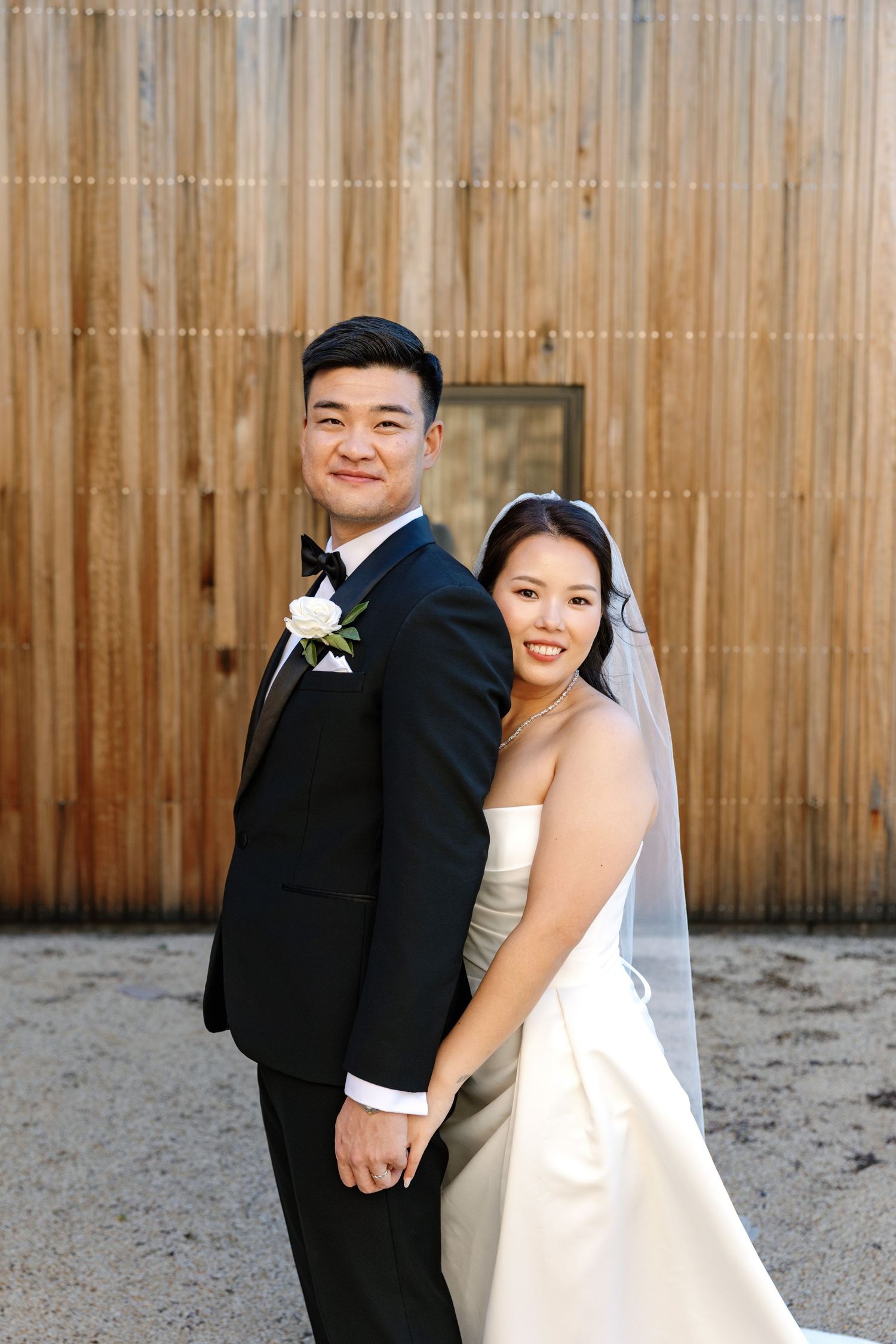 Wedding couple poses against a wooden fence backdrop in formal attire.