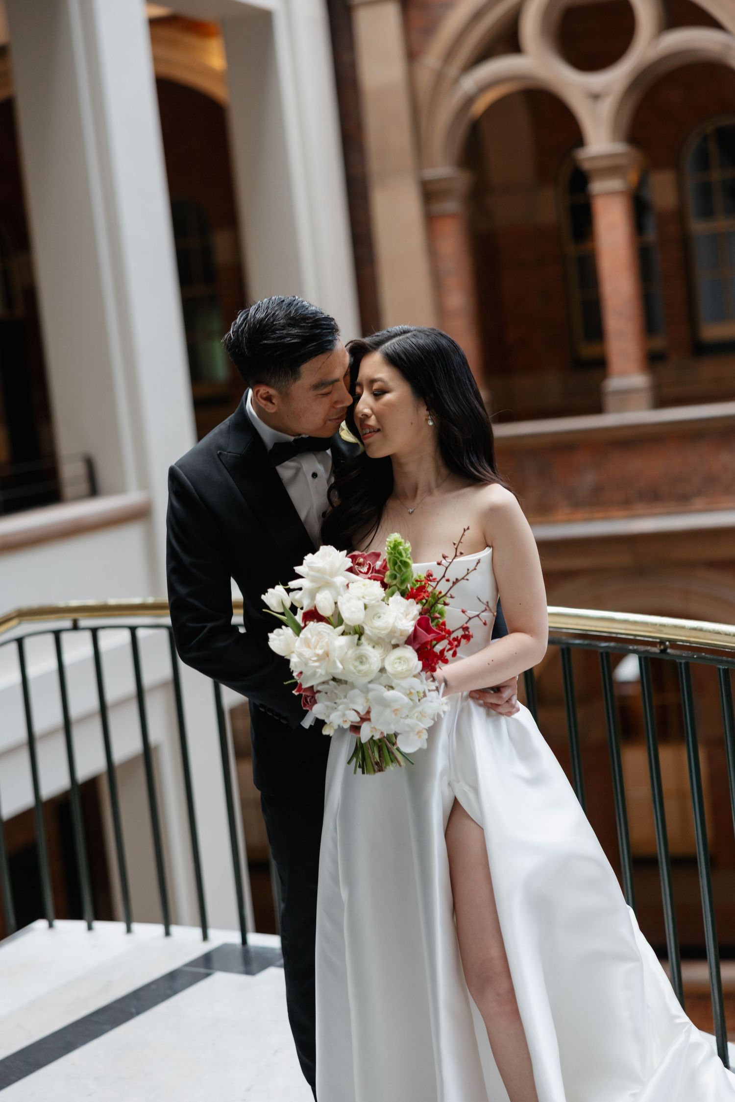 Elegant wedding couple embraces on marble staircase with white and red floral bouquet in classic architectural setting.