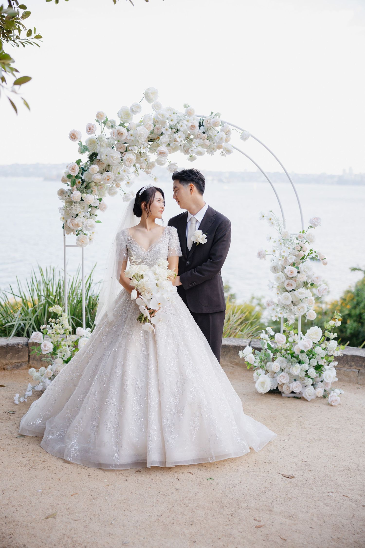 Romantic wedding couple poses under elegant white floral arch by waterfront during sunset ceremony.