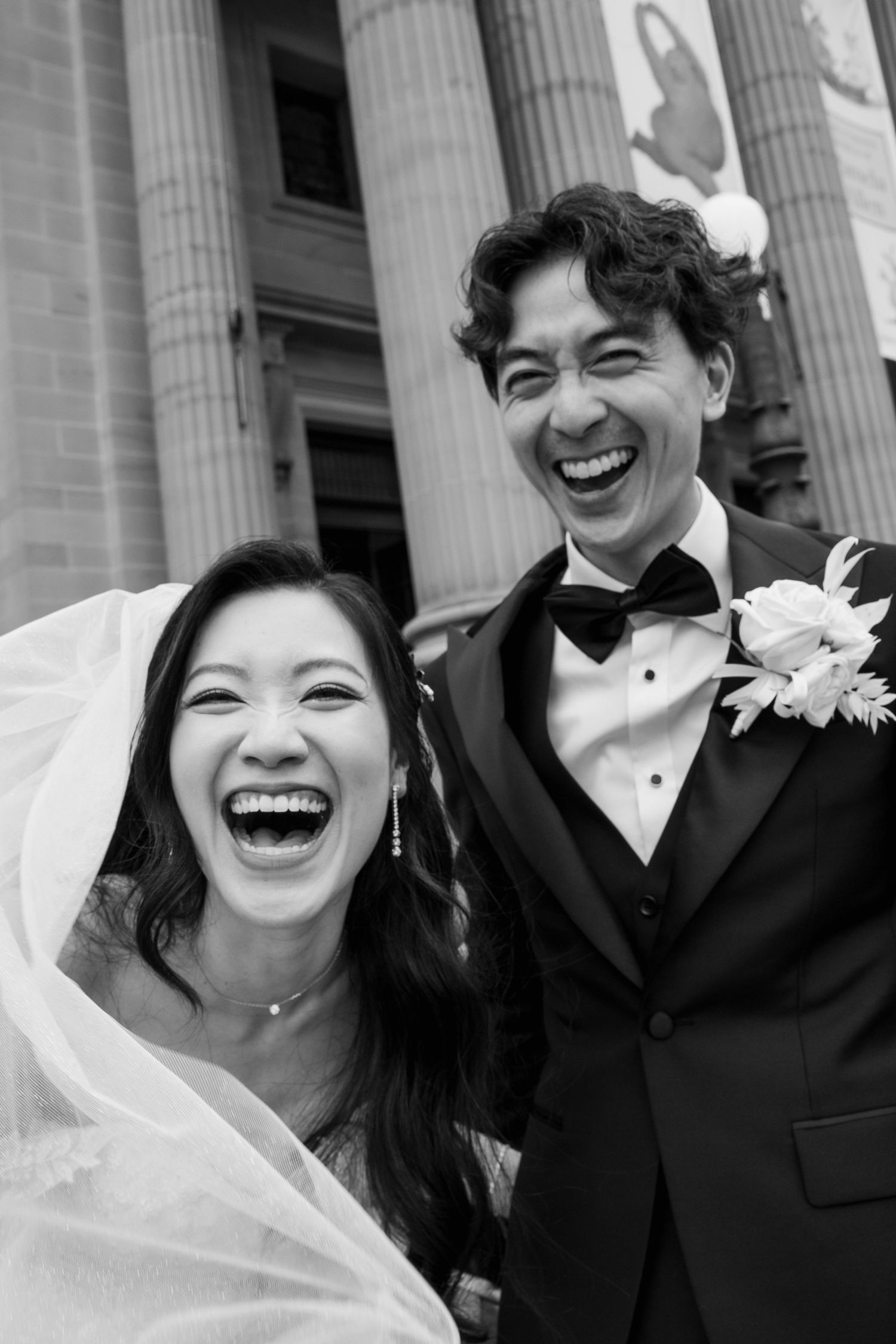 A happy bride and groom share joyful laughter in front of classic white columns on their wedding day.