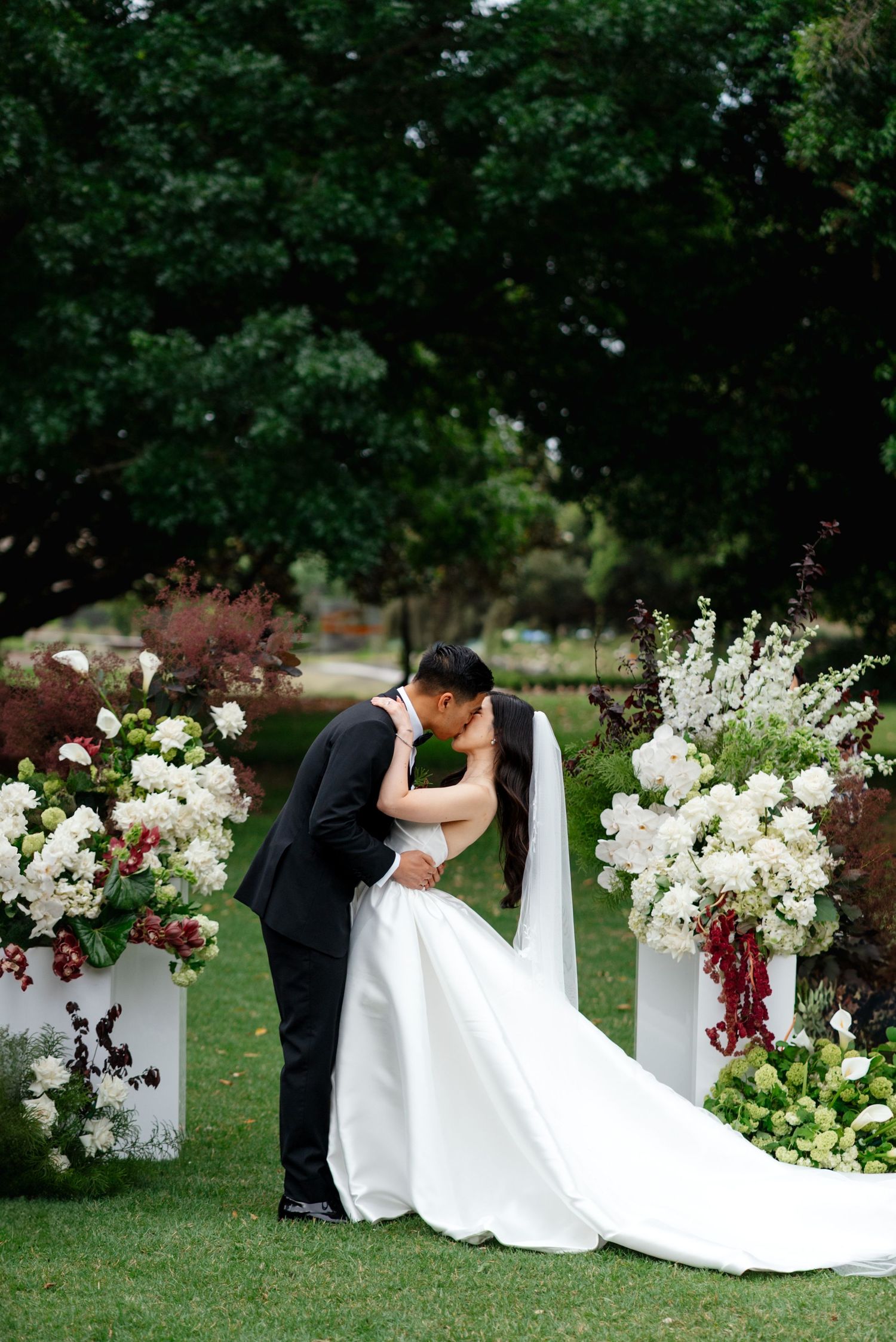 Romantic wedding couple poses among white and burgundy floral arrangements in garden setting.