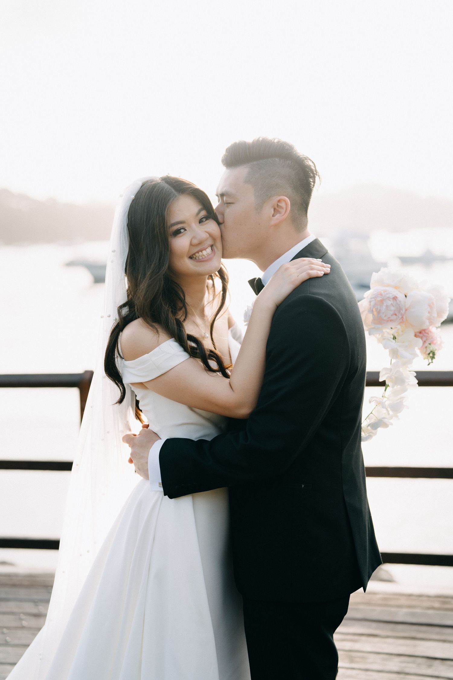 A romantic wedding couple embraces on a waterfront deck with soft lighting and floral decorations.