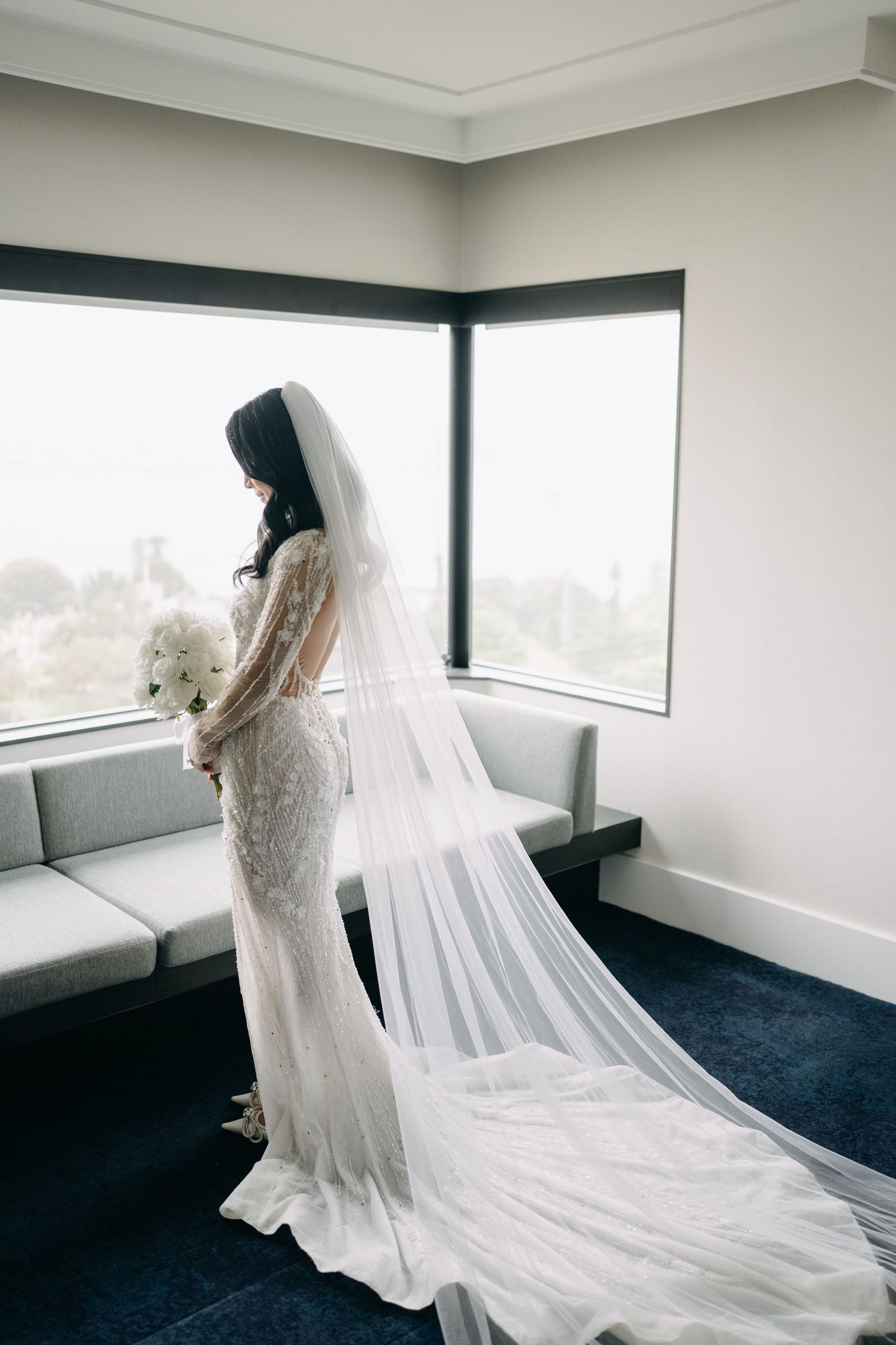 Bride in flowing lace gown with dramatic cathedral veil in modern room with windows.