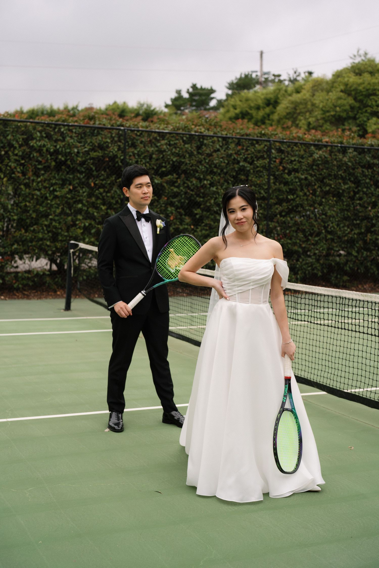 A couple in wedding attire playfully poses on a tennis court with rackets and a white dress flowing in the breeze.