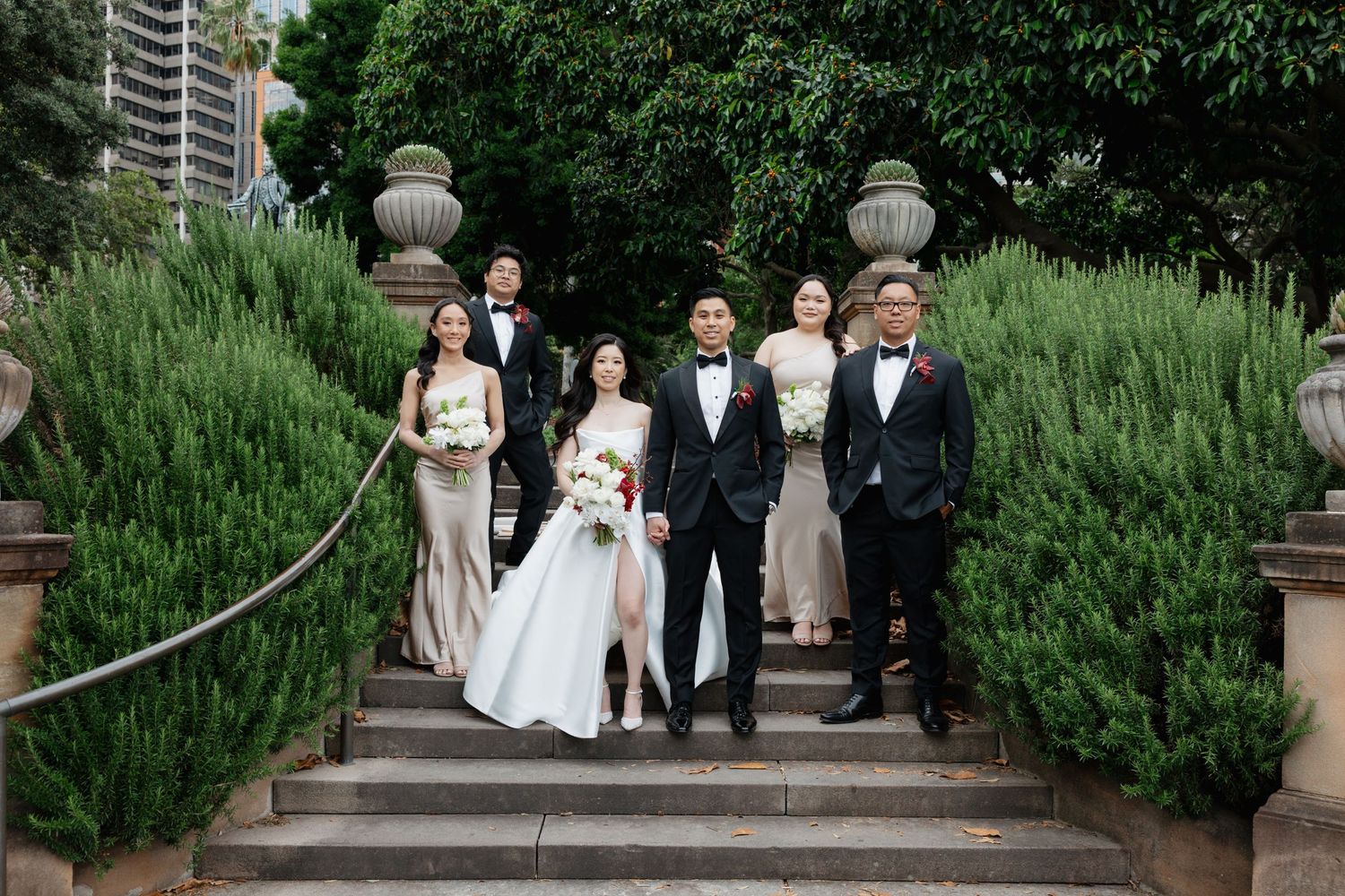 Wedding party poses on grand outdoor staircase surrounded by lush greenery at elegant venue.