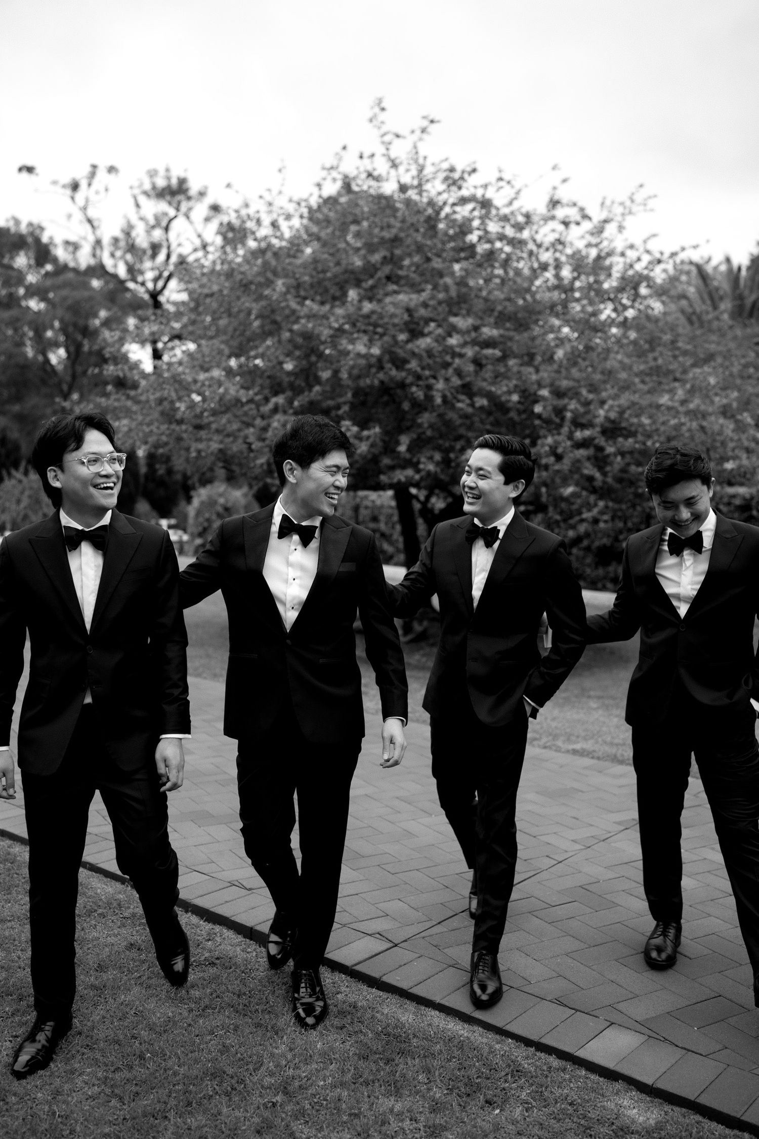 Groomsmen in black tuxedos and bow ties walk in a line outdoors.