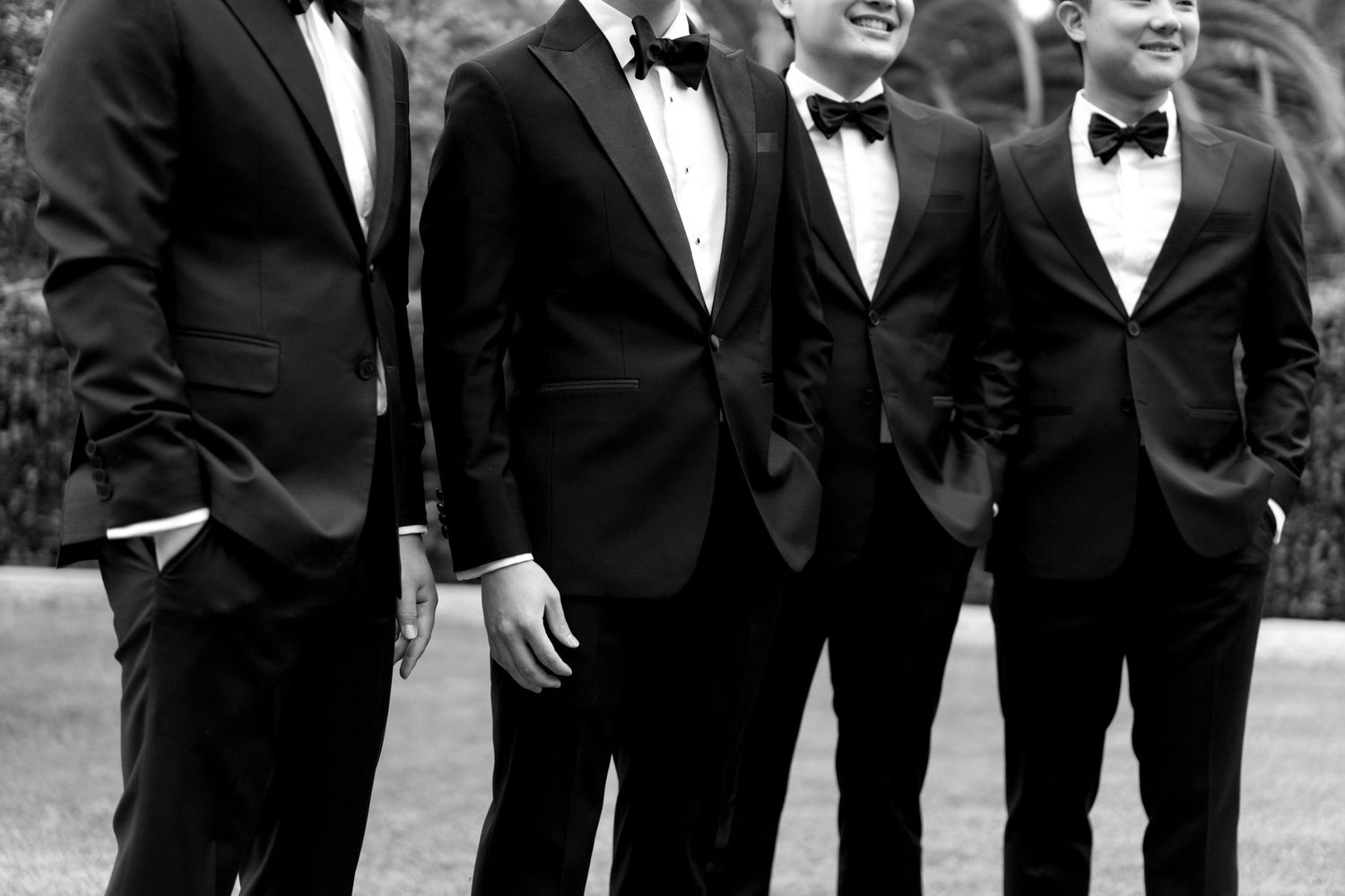A row of groomsmen in black tuxedos and bow ties stand together smiling outdoors in a black and white photo.