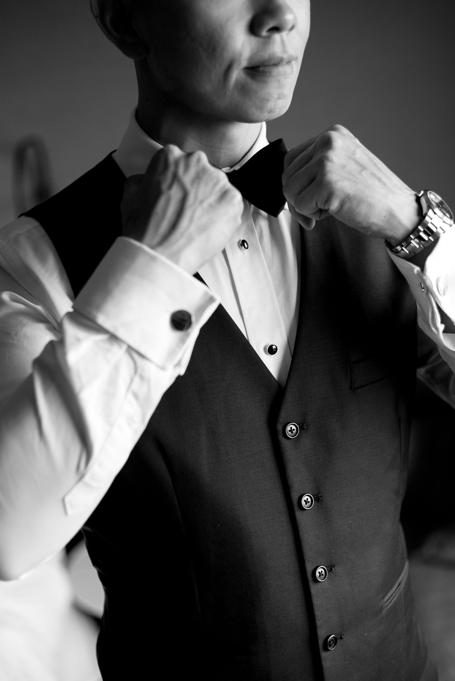 Black and white photo of someone adjusting their bowtie while wearing a formal tuxedo vest and white dress shirt.