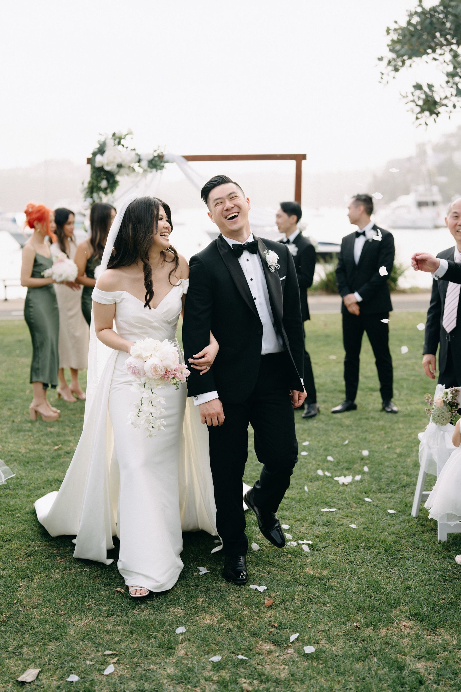 A joyful wedding couple laughs together during their outdoor ceremony with guests and floral arch in background.
