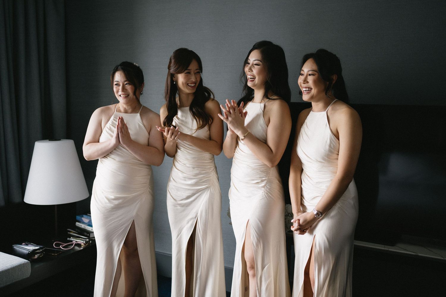 Four bridesmaids wearing elegant white floor-length dresses stand together in a dimly lit room.