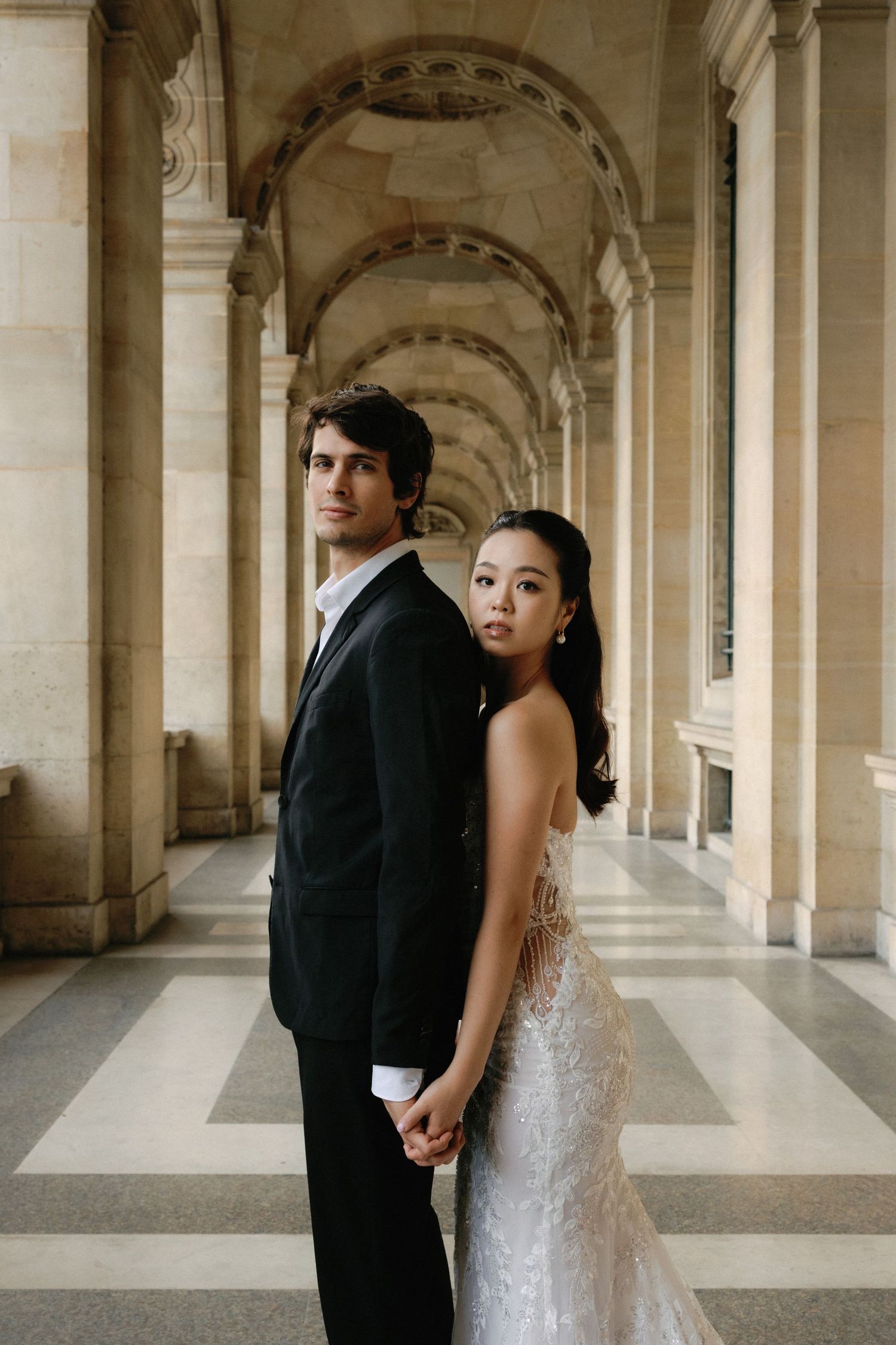 A romantic wedding portrait of a couple in formal attire standing in an ornate colonnade with arched architecture.