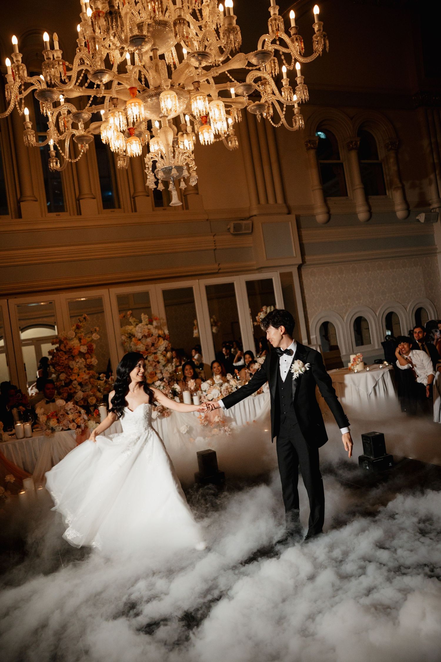 Wedding couple dances on a misty dance floor under elegant crystal chandeliers at their reception.