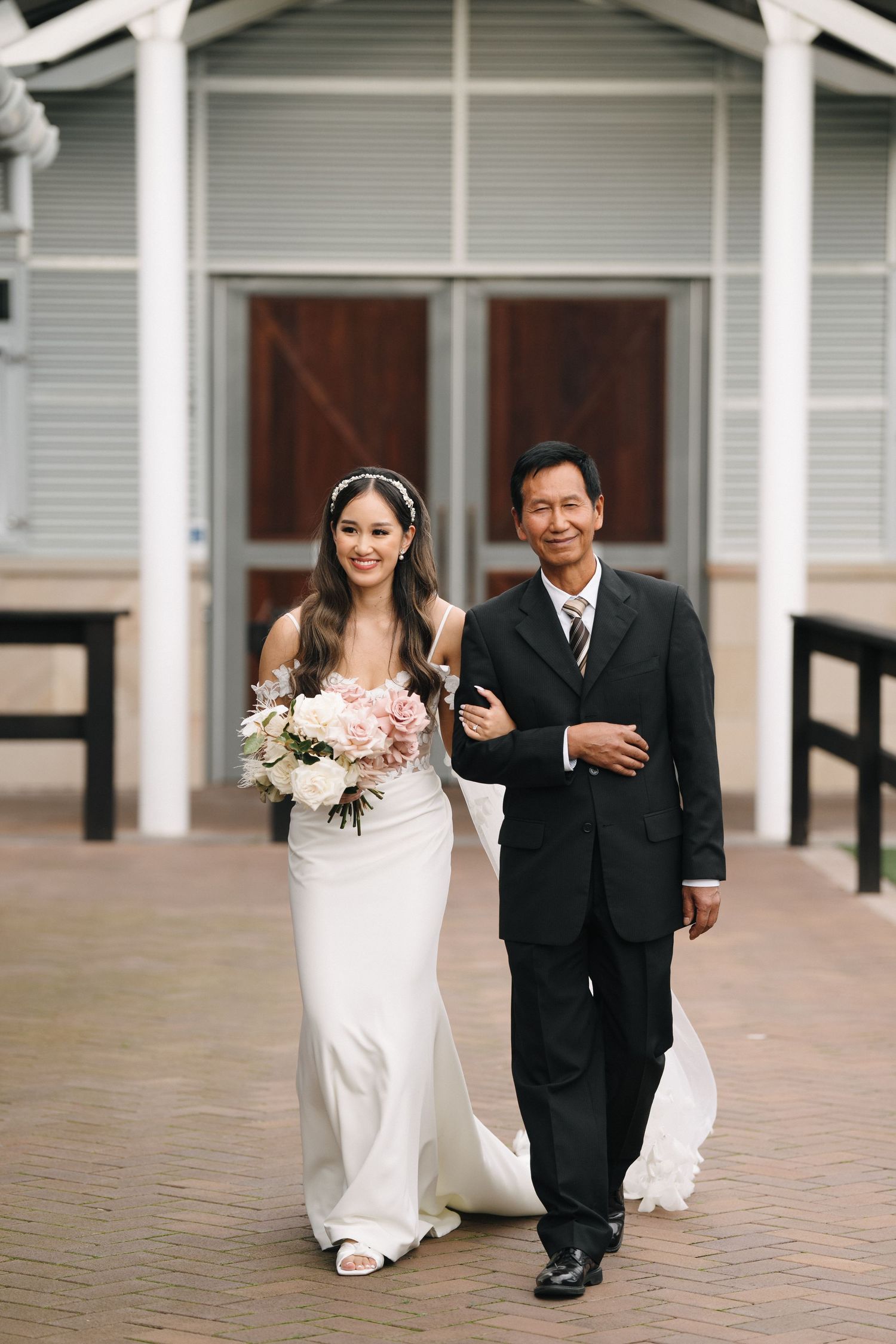 A wedding couple walks together on a brick pathway outside a building.