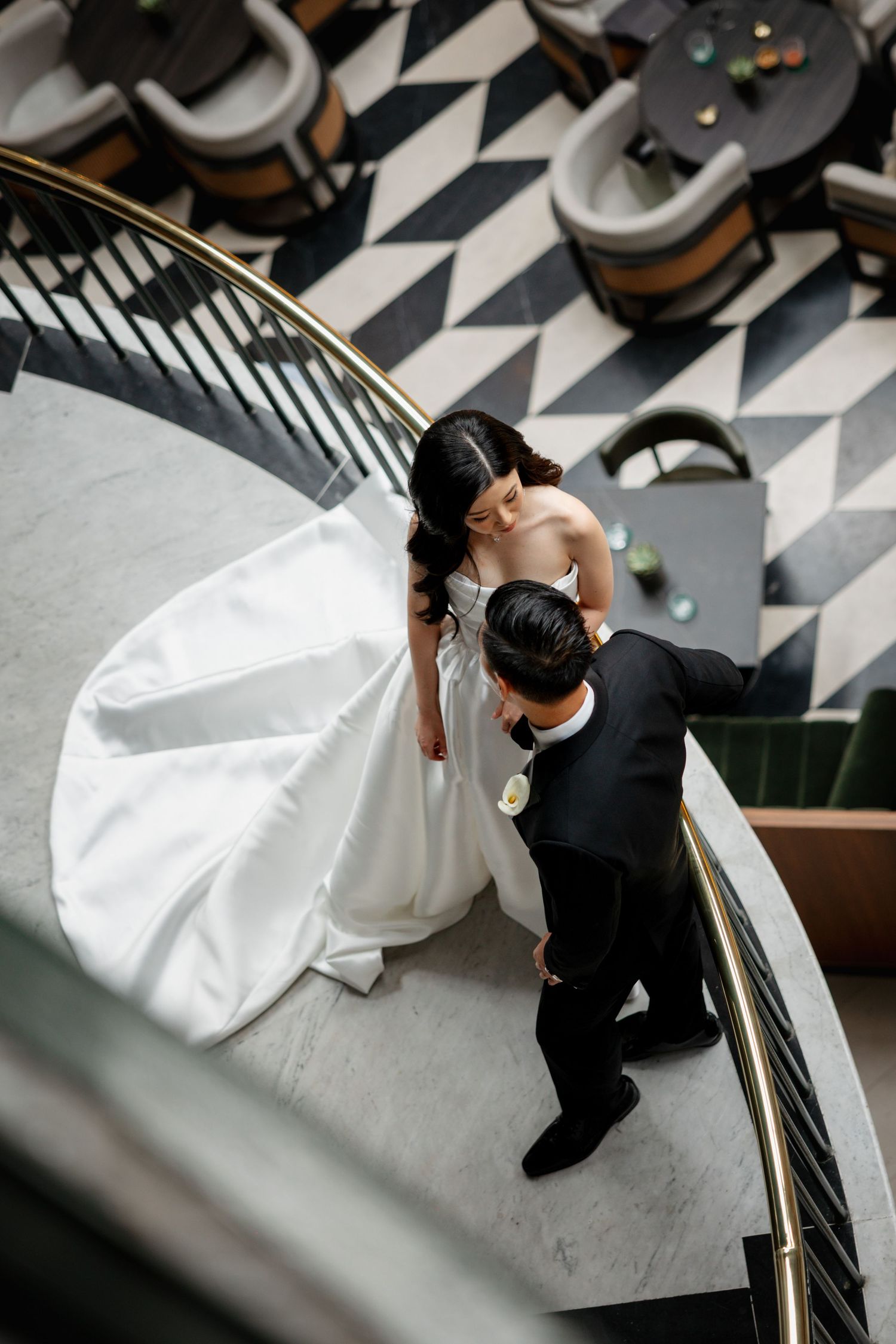 A couple in formal wedding attire descend an elegant staircase with black and white geometric flooring visible below.