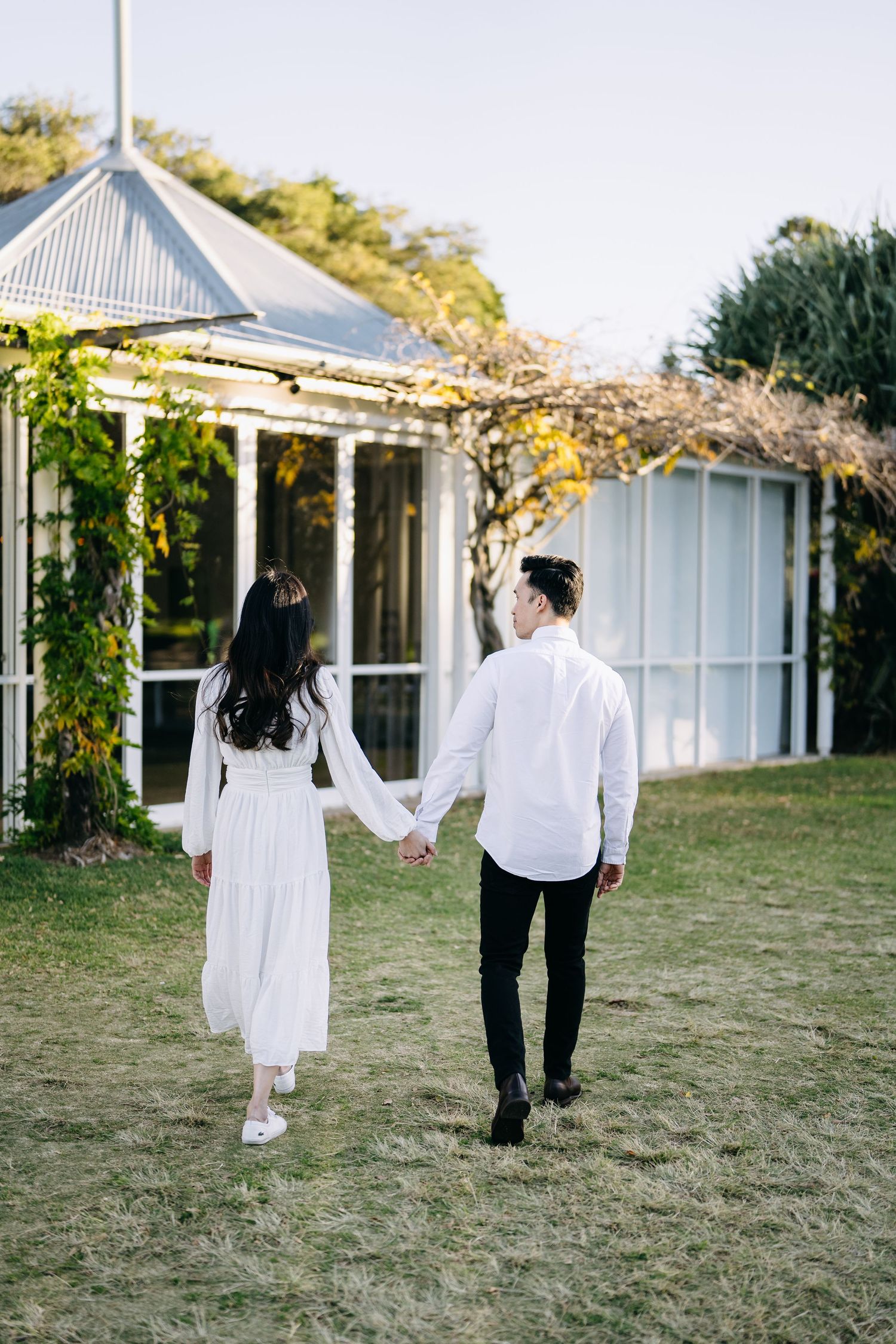 Couple walks hand in hand through garden with white cottages in background.