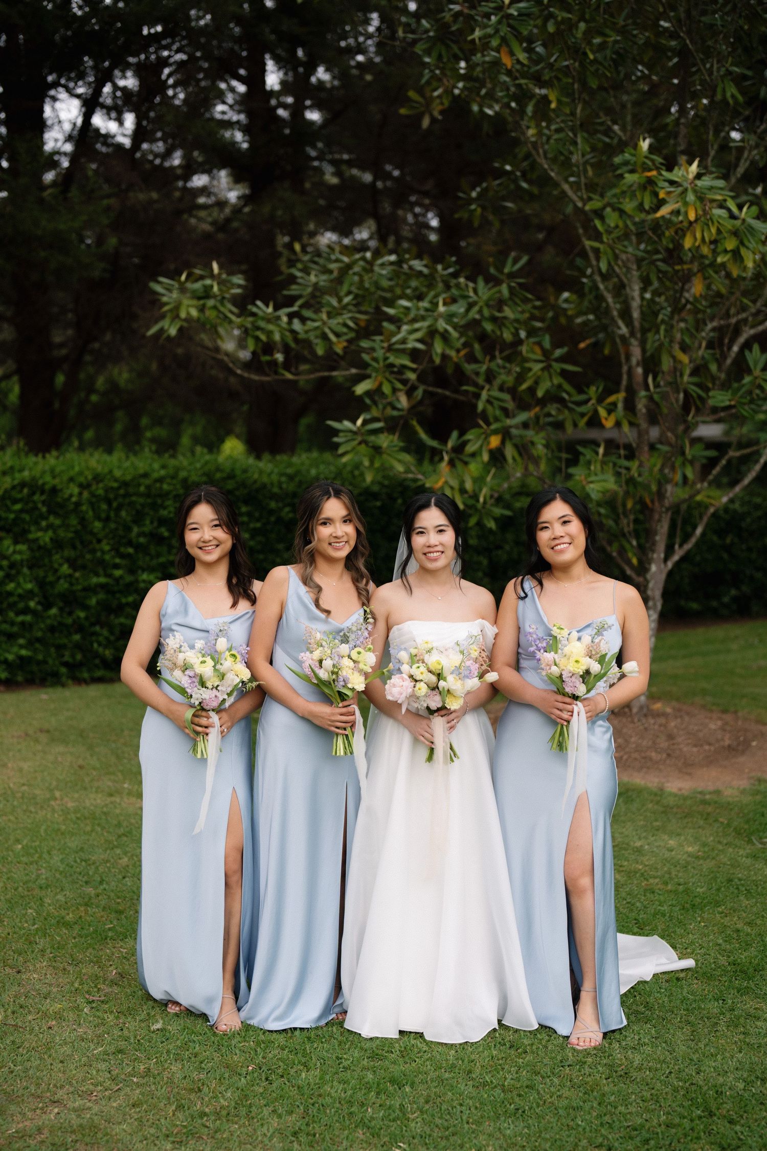 A bridal party in light blue bridesmaid dresses and white wedding gown pose together on grass with floral bouquets.