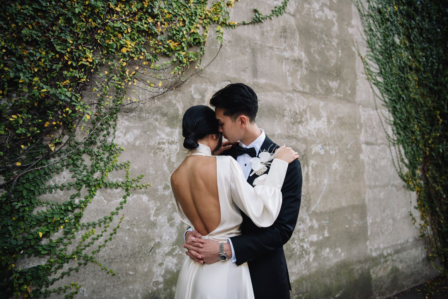 A romantic wedding couple embraces against a concrete wall with climbing ivy creating an intimate garden setting.