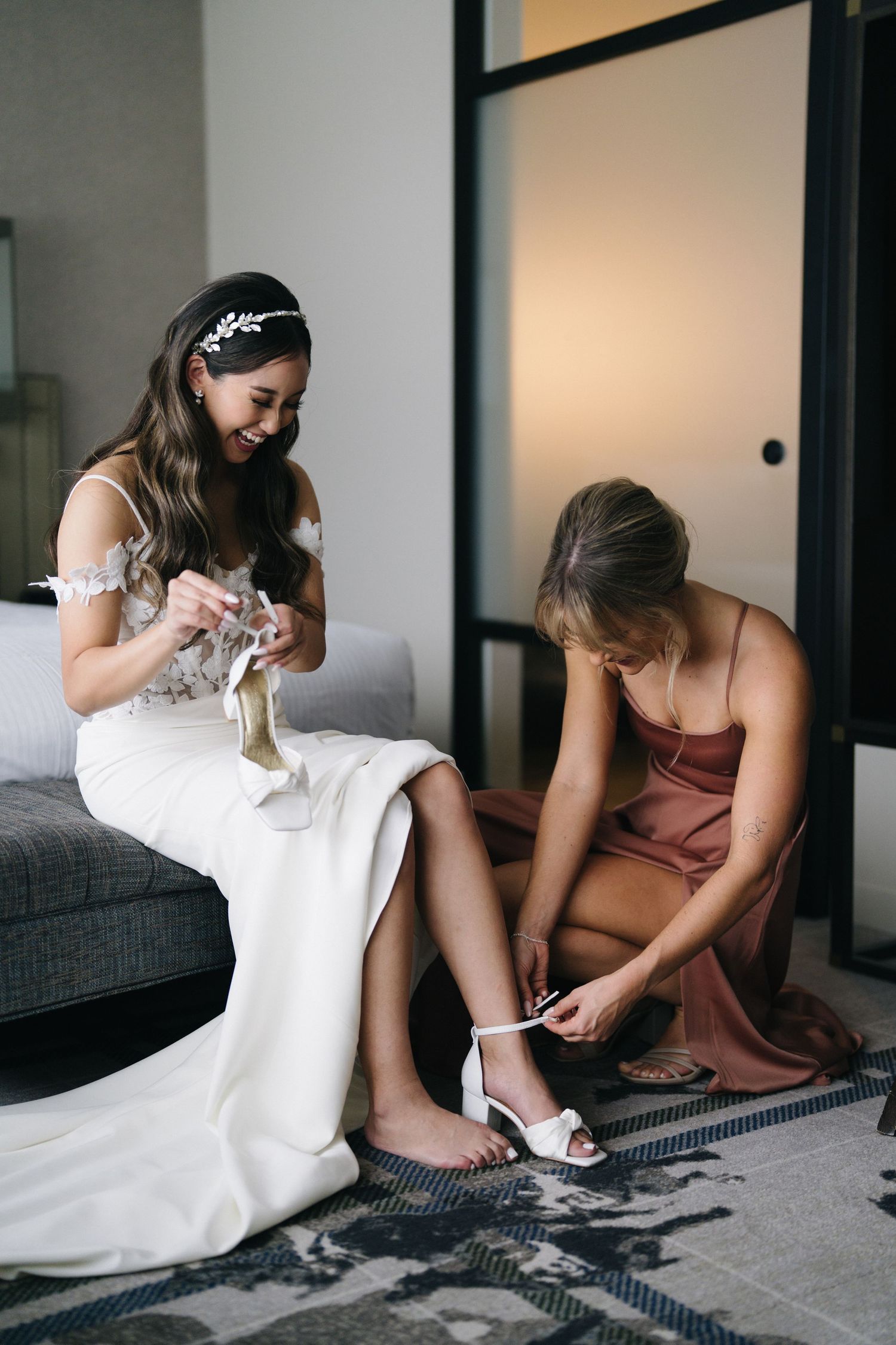 A bride getting ready in a white dress sits on a couch while someone helps with her shoes.