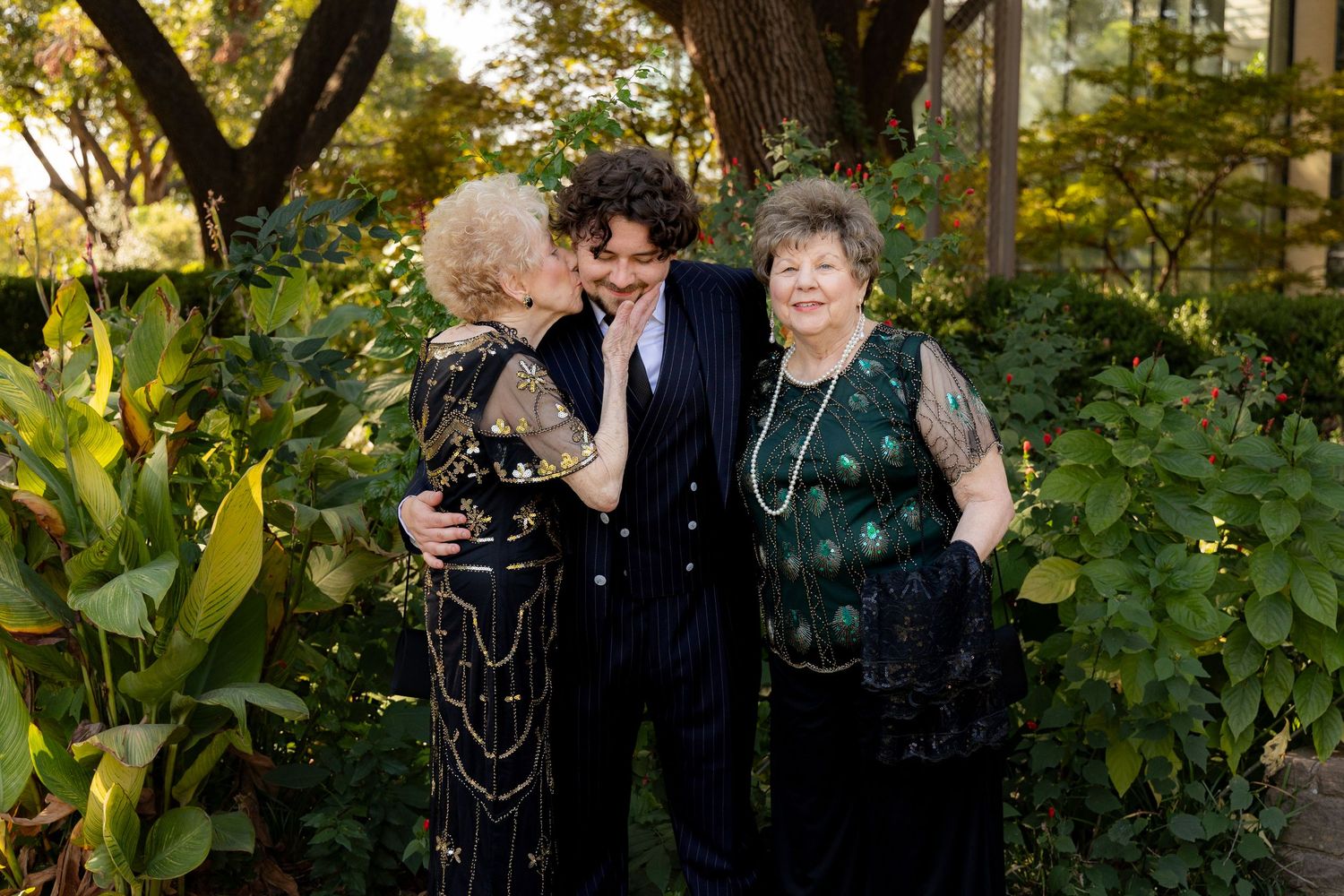 Three people in formal evening wear share a joyful moment together in a lush garden setting at sunset.