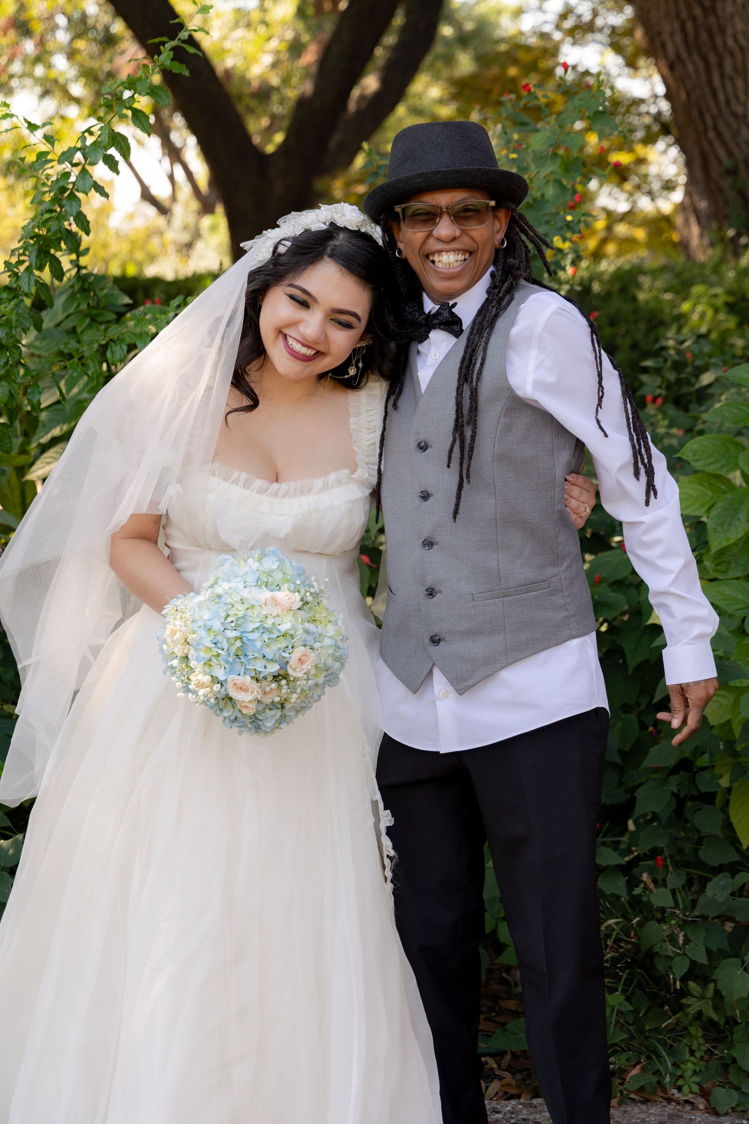Happy newlyweds pose together outdoors with bride in white gown holding blue and white bouquet.