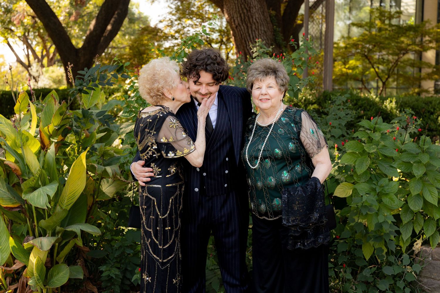 Three people dressed in formal evening wear stand together in a lush garden setting during golden hour.