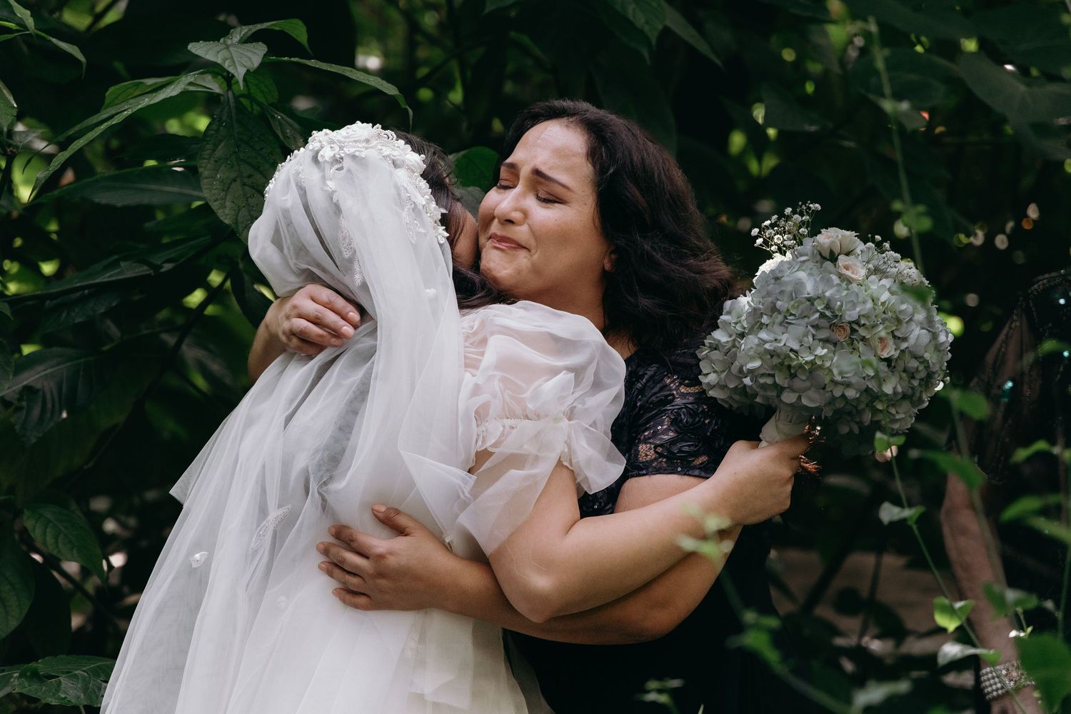 Someone embraces a wedding dress and holds a bouquet while standing in front of green foliage outdoors.