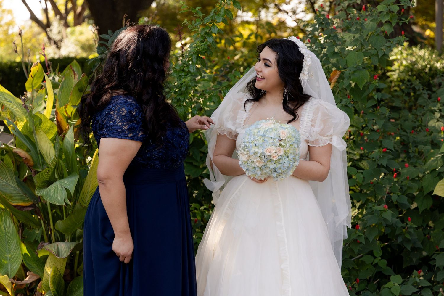 Two figures in a garden setting, one wearing a white wedding dress with a veil and another in a navy blue dress.