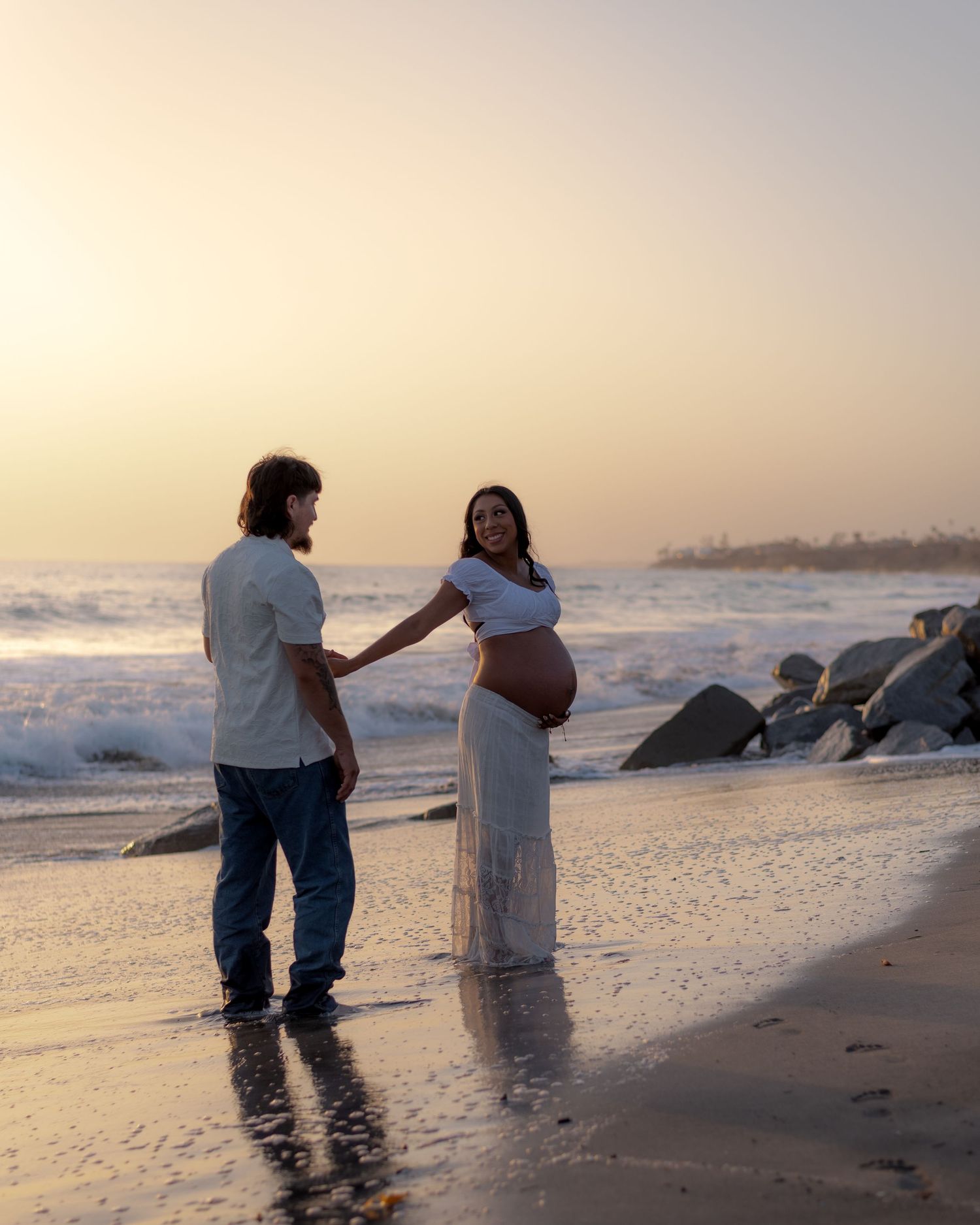 Pregnant woman in white dress holding partner's hand on reflective beach at golden sunset.